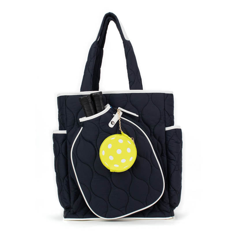 The Queen of the Court Fifth Ave Pickleball Convertible Tote Bag/Backpack in navy blue with white trim features outer pockets. Two black-handled paddles fit in a front pocket, and a round yellow "Queen of the Court" accessory hangs nearby.