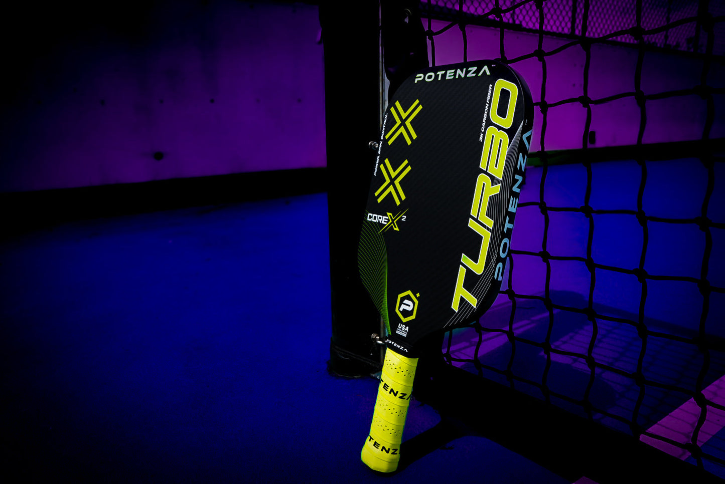 A Potenza Pickleball TURBO+ COREx2 carbon fiber paddle with neon yellow accents and bright yellow handle grip rests against a black net post on a blue court, illuminated by dramatic purple lighting in the background.