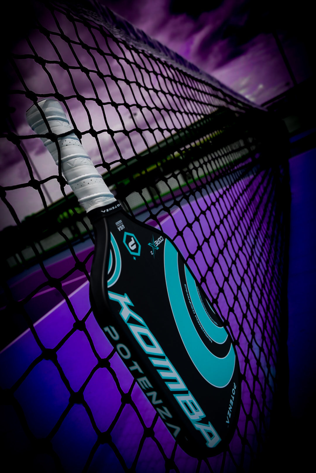 A black and turquoise carbon fiber Potenza Pickleball KOMBA+ COREx2 paddle leans against a net on a court beneath a dramatic purple sky, with striking shadows and vivid colors enhancing the energetic, moody scene.