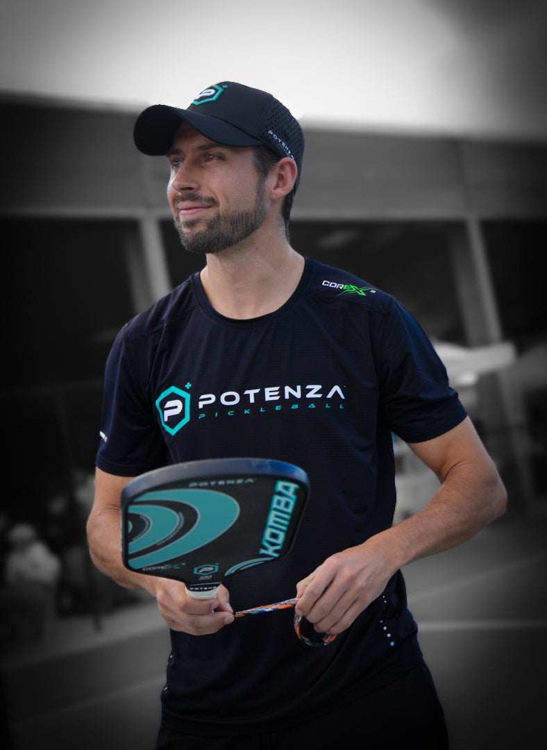 A man with a trimmed beard, wearing a black Potenza Pickleball Quick-Dry Performance T-Shirt and cap, stands on a court holding a green pickleball paddle. The blurred grayscale background draws focus to him as he gazes aside with a slight smile.