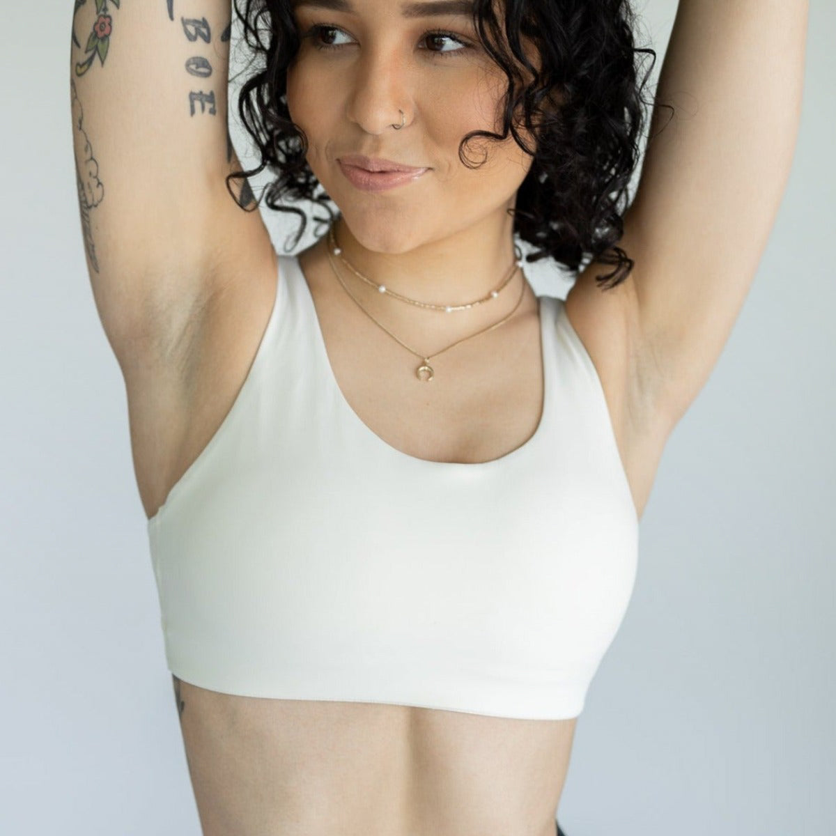 A person with curly dark hair, tattoos, and a nose ring wears the DYM Athletics Element Essential Bra (A-D cup, medium support) with adjustable straps. They smile softly, raising both arms and looking sideways against a light background.