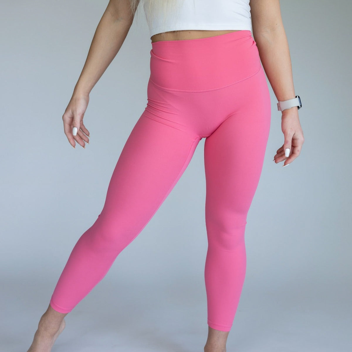 Against a plain background, a person wears DYM Athletics' Ultra High Waist Flex & Flow Legging in bright pink with a white crop top. Their face is out of frame, long blond hair visible, and they wear a white smartwatch on their left wrist.