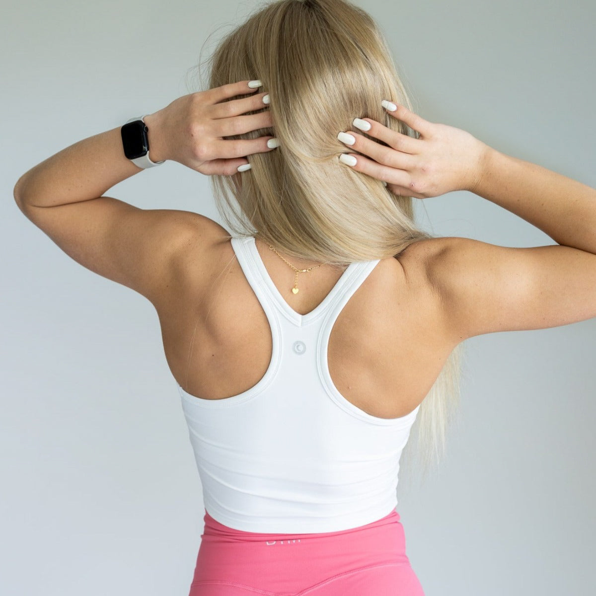 A woman with long blonde hair is seen from behind wearing a white DYM Athletics Energy Crop Everyday Tank and pink leggings. She has manicured nails, a smartwatch on her left wrist, and a delicate necklace, with both hands raised to her head.