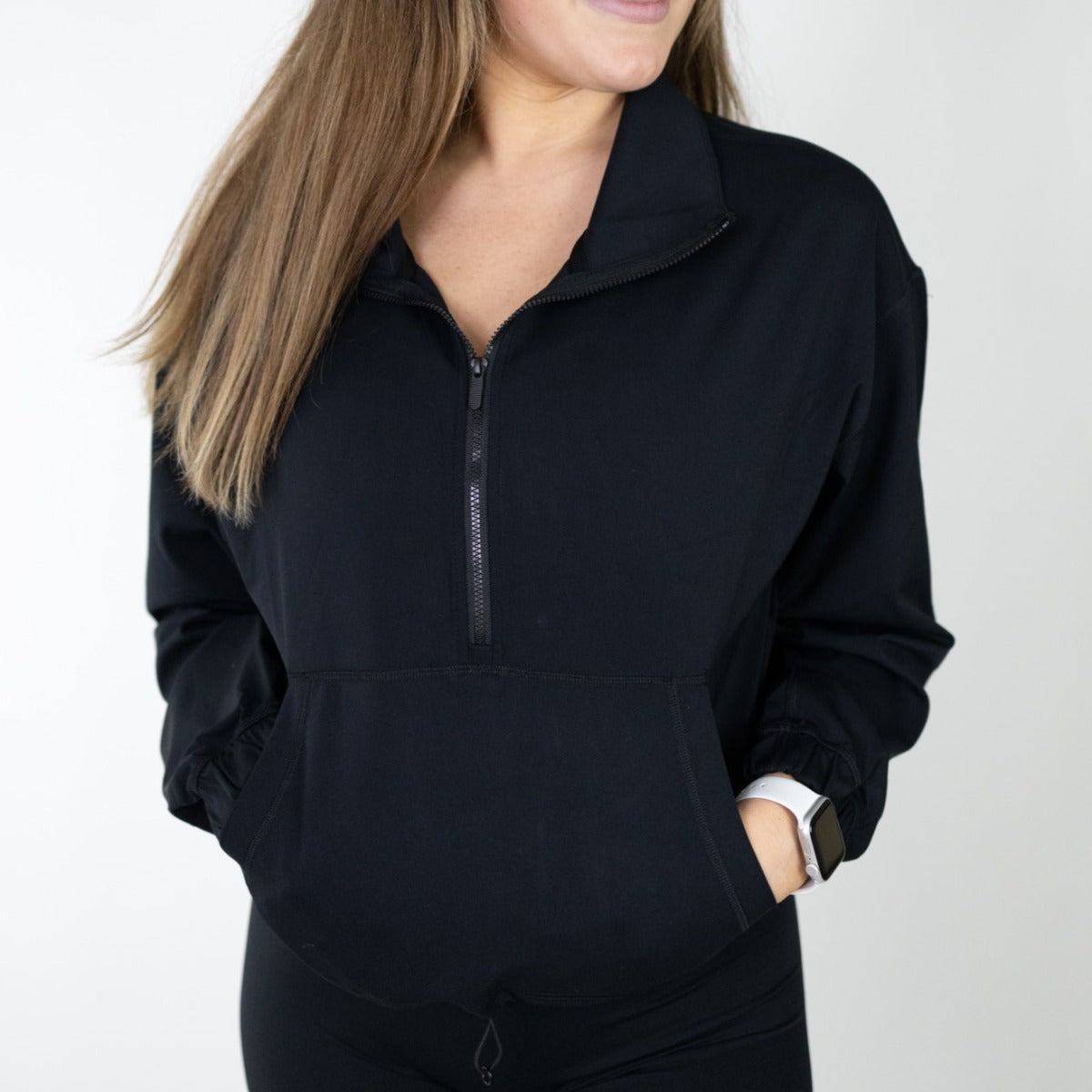 A woman with long, straight light brown hair wears the DYM Athletics Ascend 1/4 Zip Jacket in black. She stands against a plain white background with her hands in the front pocket and a white smartwatch on her left wrist; face partially out of frame.
