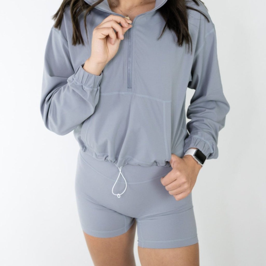 A woman showcases her smartwatch while wearing DYM Athletics’ Serene Biker Short | 5" | SALE in light gray, paired with a matching half-zip long-sleeve top. She stands against a white background; her head is not visible.