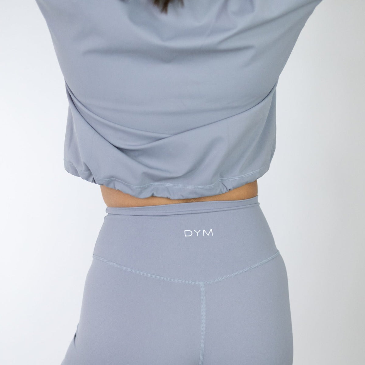 A person wears a light gray crop top and high-waisted Serene Biker Shorts (5", DYM Athletics) with a seamless front and "DYM" on the waistband. The background is plain white.