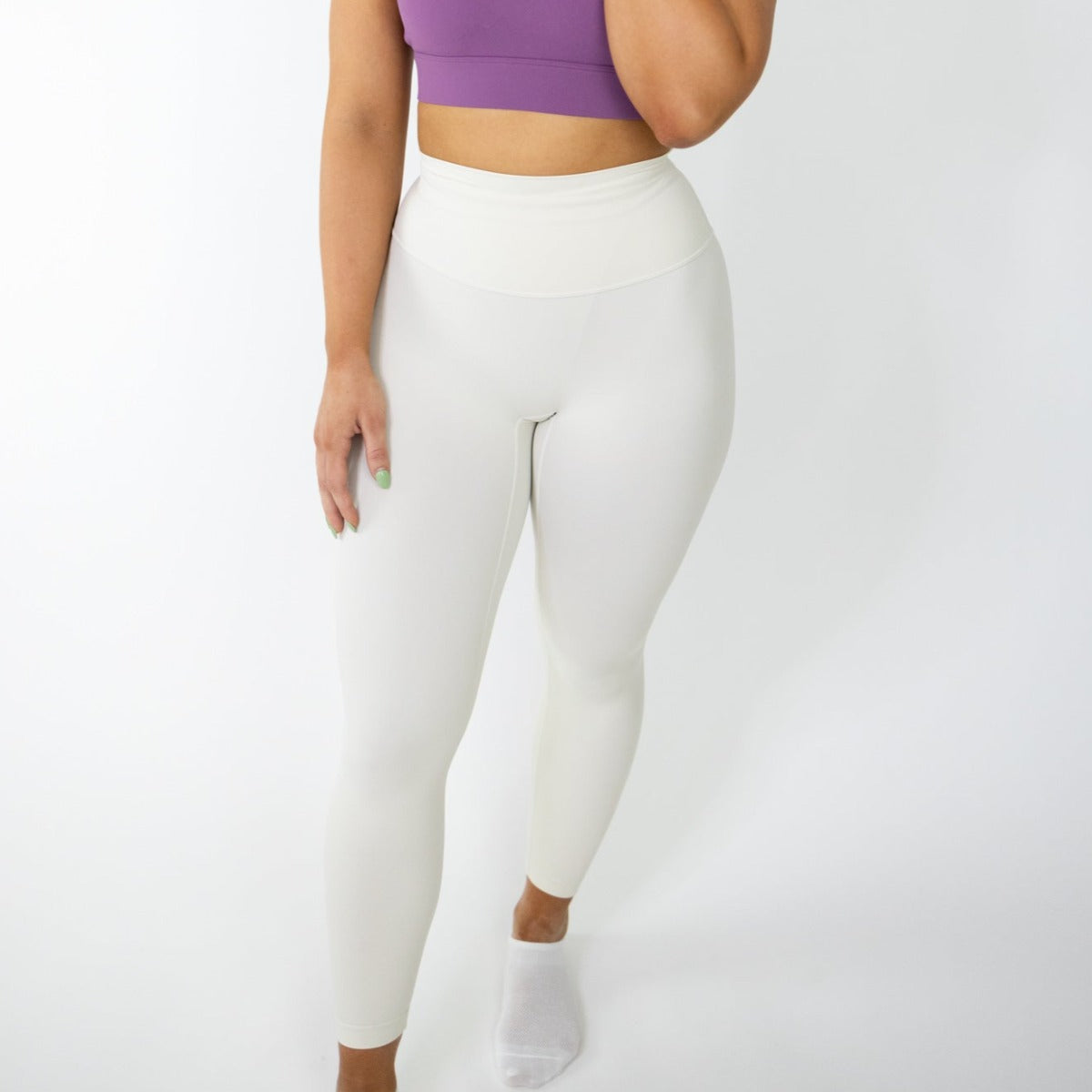A woman with light skin wears DYM Athletics’ Seamlessly Free Legging in white, paired with a purple crop top. She stands against a white background, face unseen, touching her chin. She also has white ankle socks and light green nails.