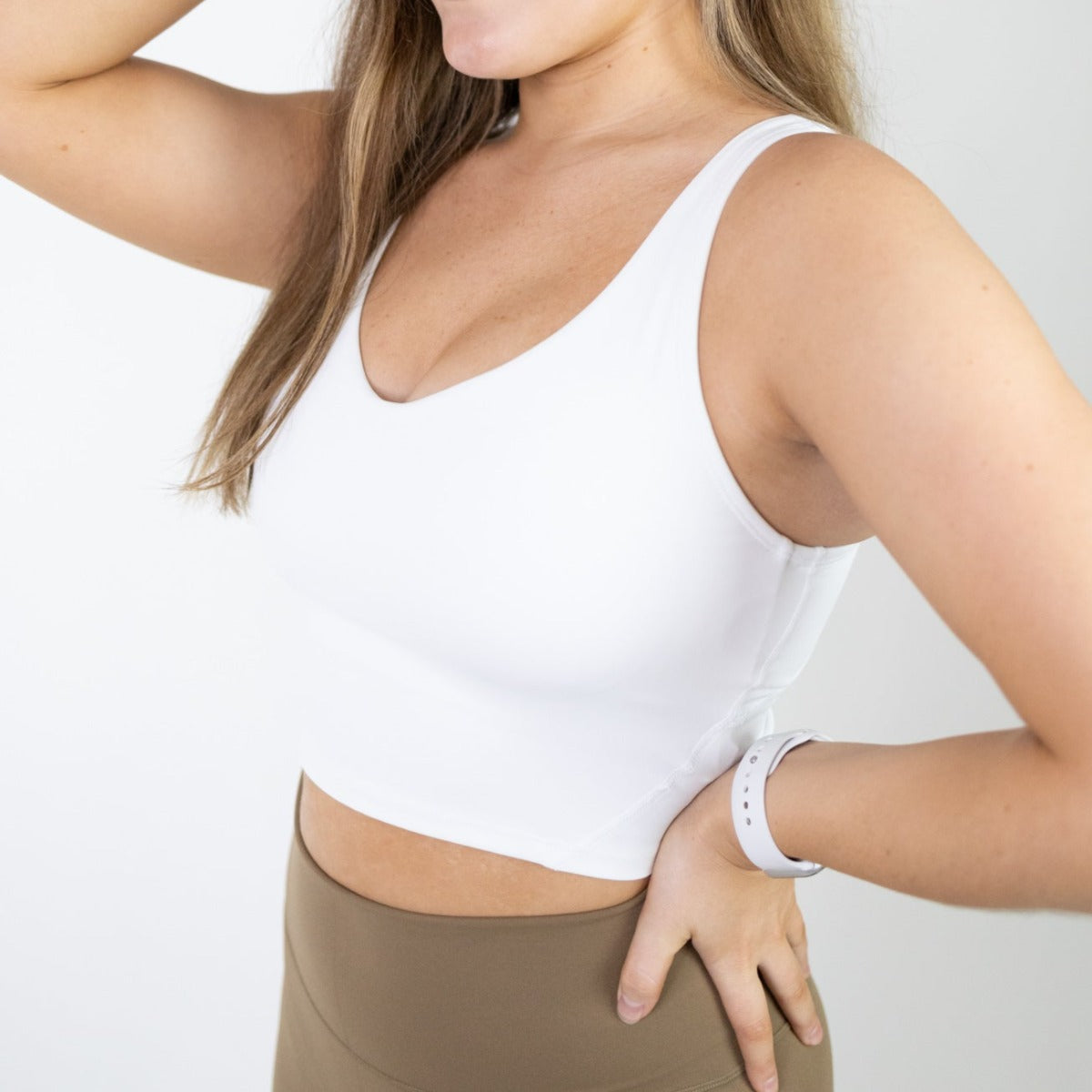 A woman poses against a white background, wearing the DYM Athletics Flex V-Neck Bra with Scoop Back in brown high-waisted leggings and a white smartwatch. Her long, light brown hair is down and she has one hand on her hip.