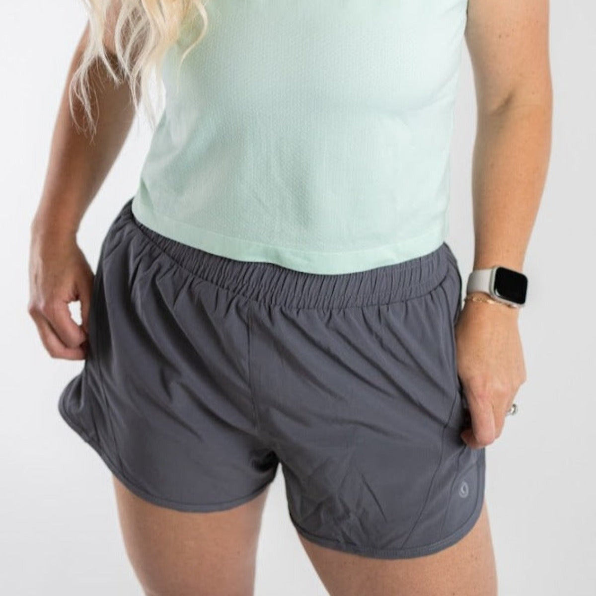 A person with long, wavy blonde hair stands against a plain white background, hands in the zipper pockets of DYM Athletics Speed Mid-Rise Shorts | 2", paired with a light mint green t-shirt, silver smartwatch, and a ring on their left hand.