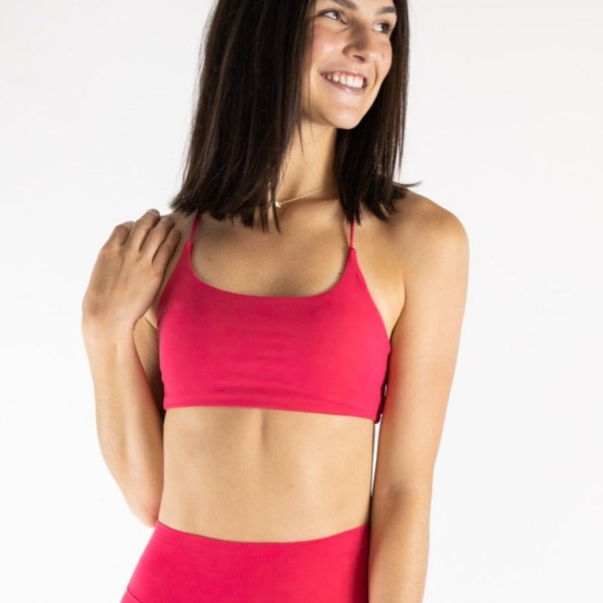 A woman with straight, shoulder-length dark hair smiles and looks left, wearing the DYM Athletics AIRE Ultra-Soft Open Back Bra in bright pink. Her right hand touches her hair as she poses against a white background in matching leggings.