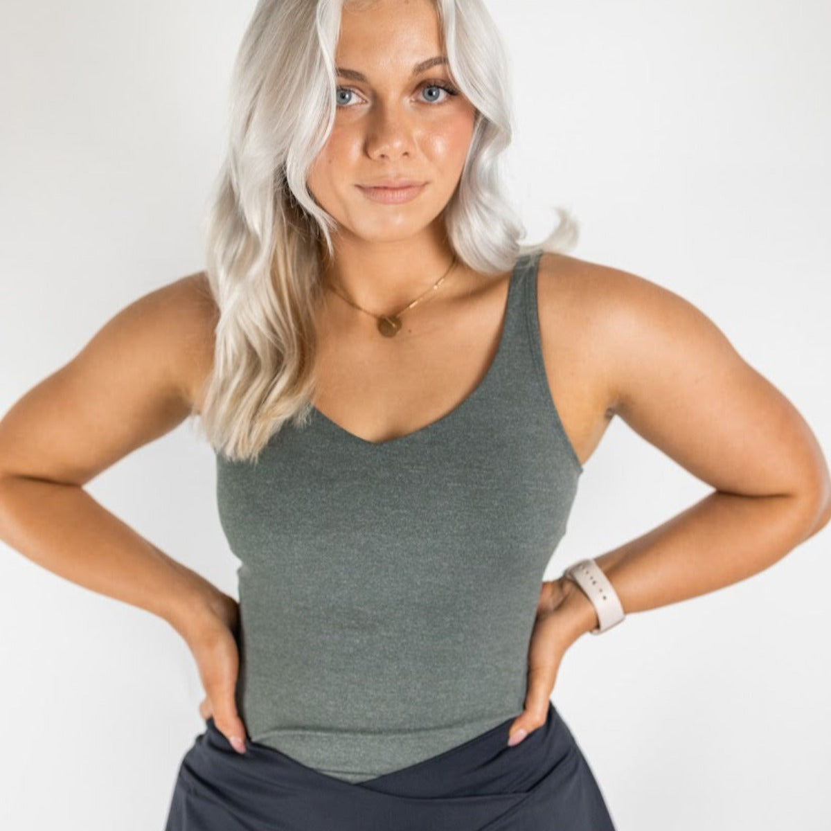 A woman with long, wavy, platinum blonde hair stands against a white background wearing the DYM Athletics Bliss"Full" V-Neck Tank with Scoop Back (A-D Cup), a gold necklace, and a light smartwatch, hands on hips as she looks confidently at the camera.