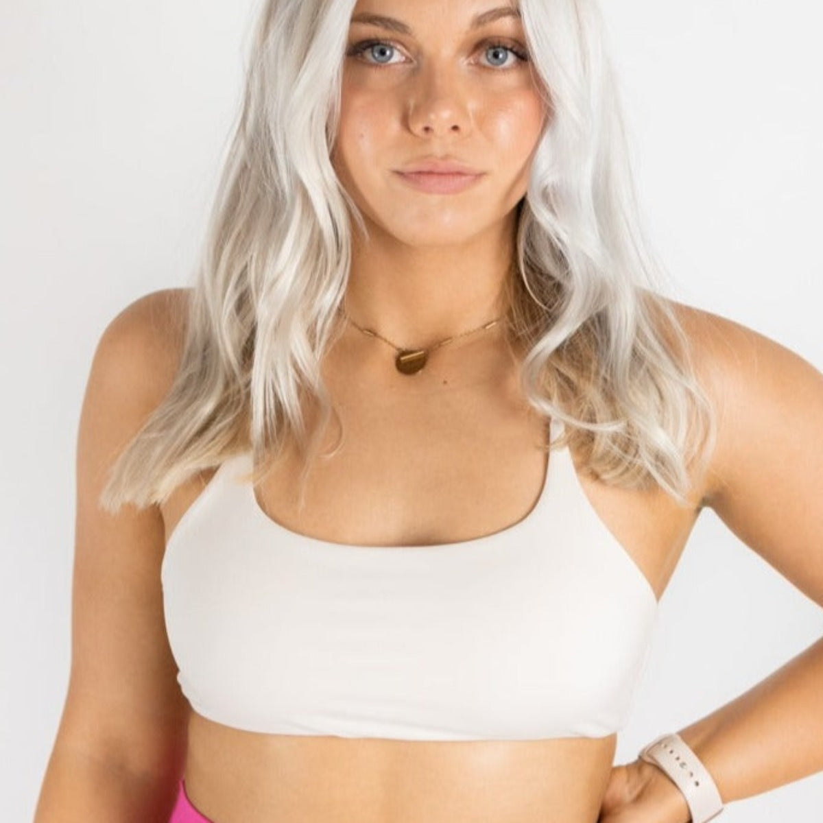 A young woman with long, wavy platinum blonde hair wears the DYM Athletics AIRE Ultra-Soft Open Back Bra (A-C Cup, Light Support), pink bottoms, a gold heart necklace, and a beige smartwatch, standing against a white background with a neutral expression.