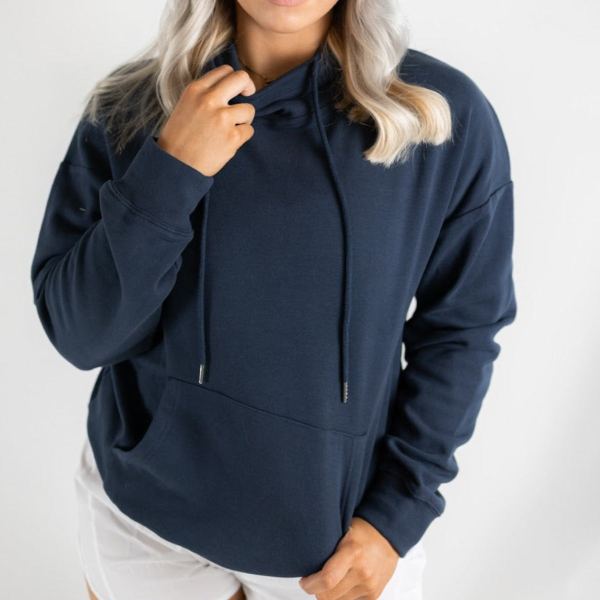 A person with wavy blond hair wears the DYM Athletics Cozy Up Cowl Neck Hoodie in dark blue with drawstrings and a front pocket, paired with white shorts. Standing against a light background, they lightly hold the hoodie near their neck.