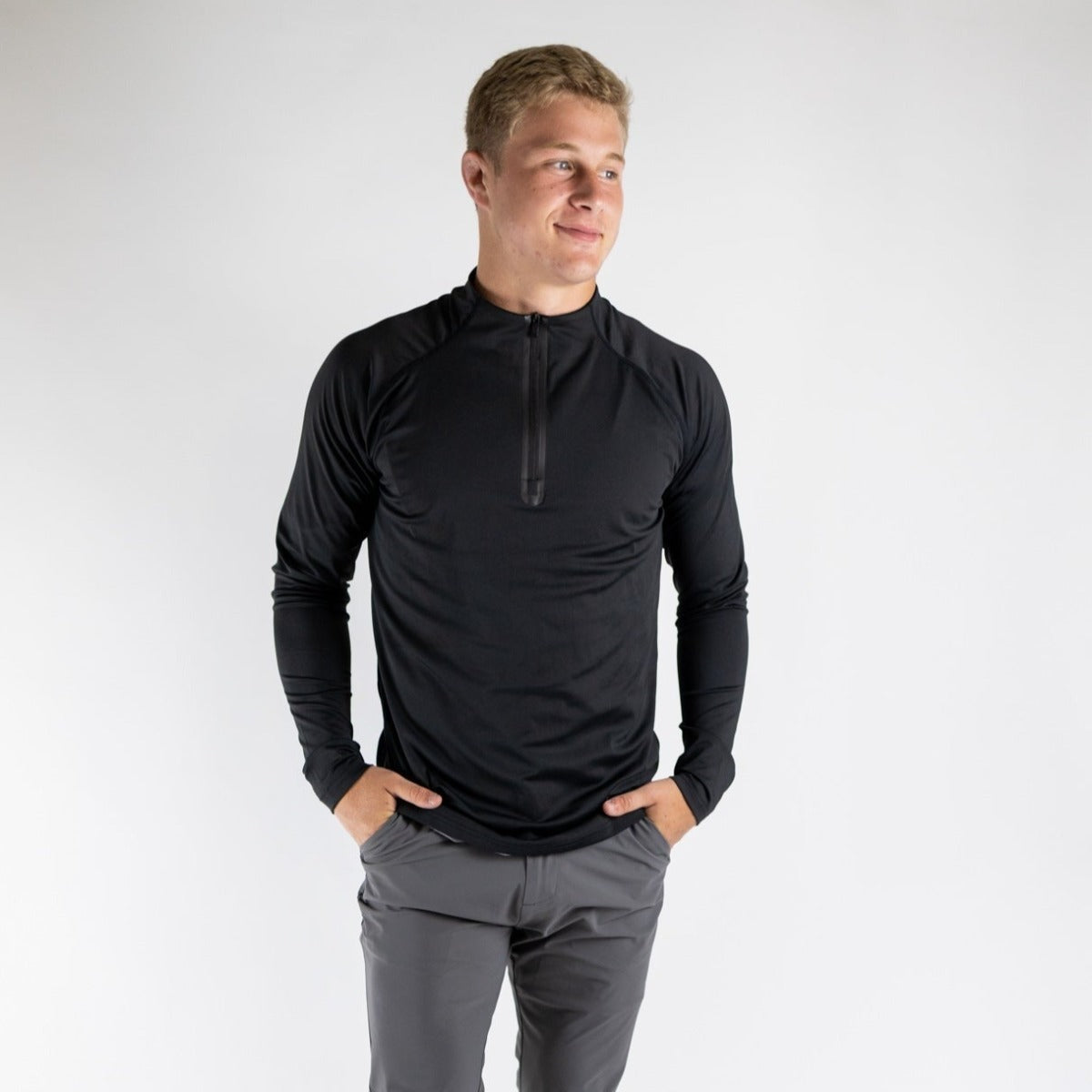A young man with short blond hair smiles slightly, standing against a white background. He wears the DYM Athletics Top-Notch Men's 1/4 Zip in black and gray golf shorts—perfect activewear on SALE. Hands in pockets, he’s ready for anything.