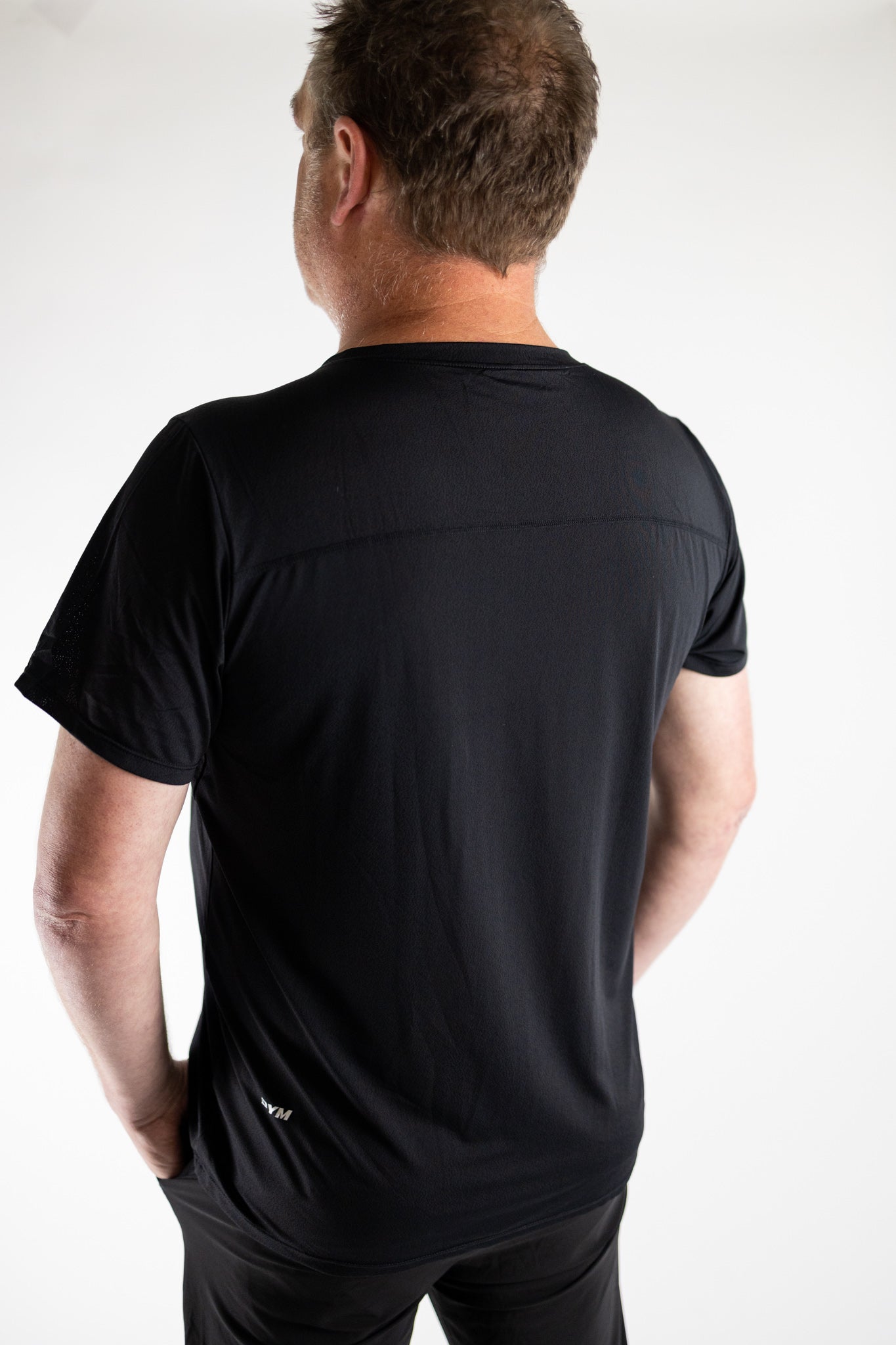 A man with short, tousled light brown hair stands with his back to the camera against a white background, wearing DYM Athletics' Elevate Men's Lightweight Breathable Tee in black and matching black pants.