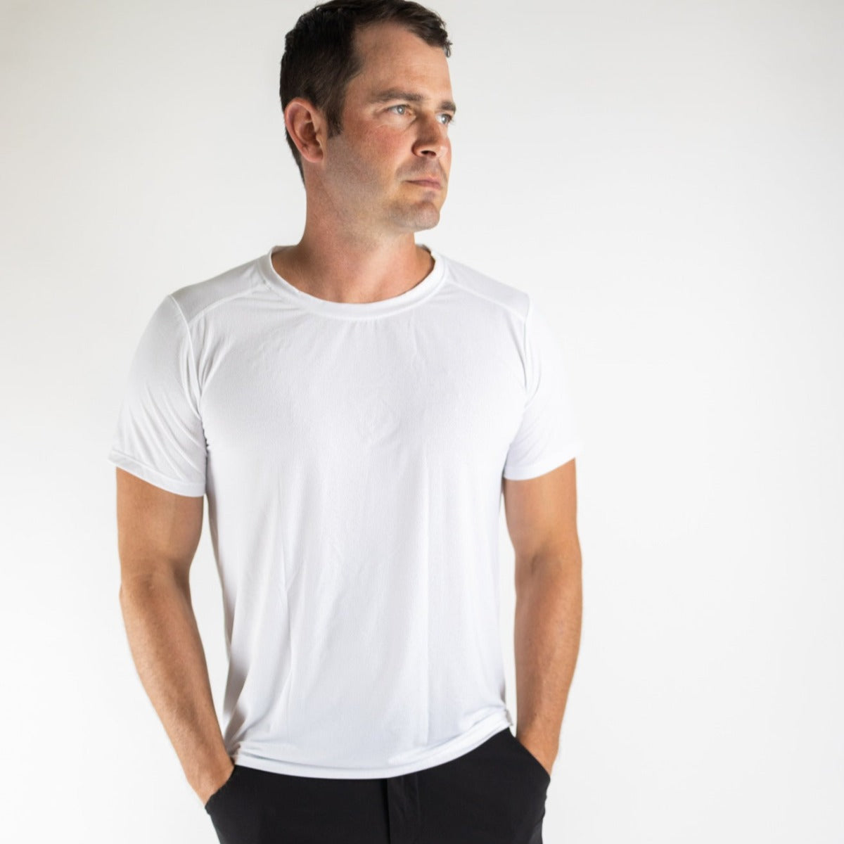 A man with short dark hair stands against a white background wearing the DYM Athletics Elevate Men's Lightweight Breathable Tee (SALE) and black pants, his hands in his pockets as he looks off to the side with a neutral expression.