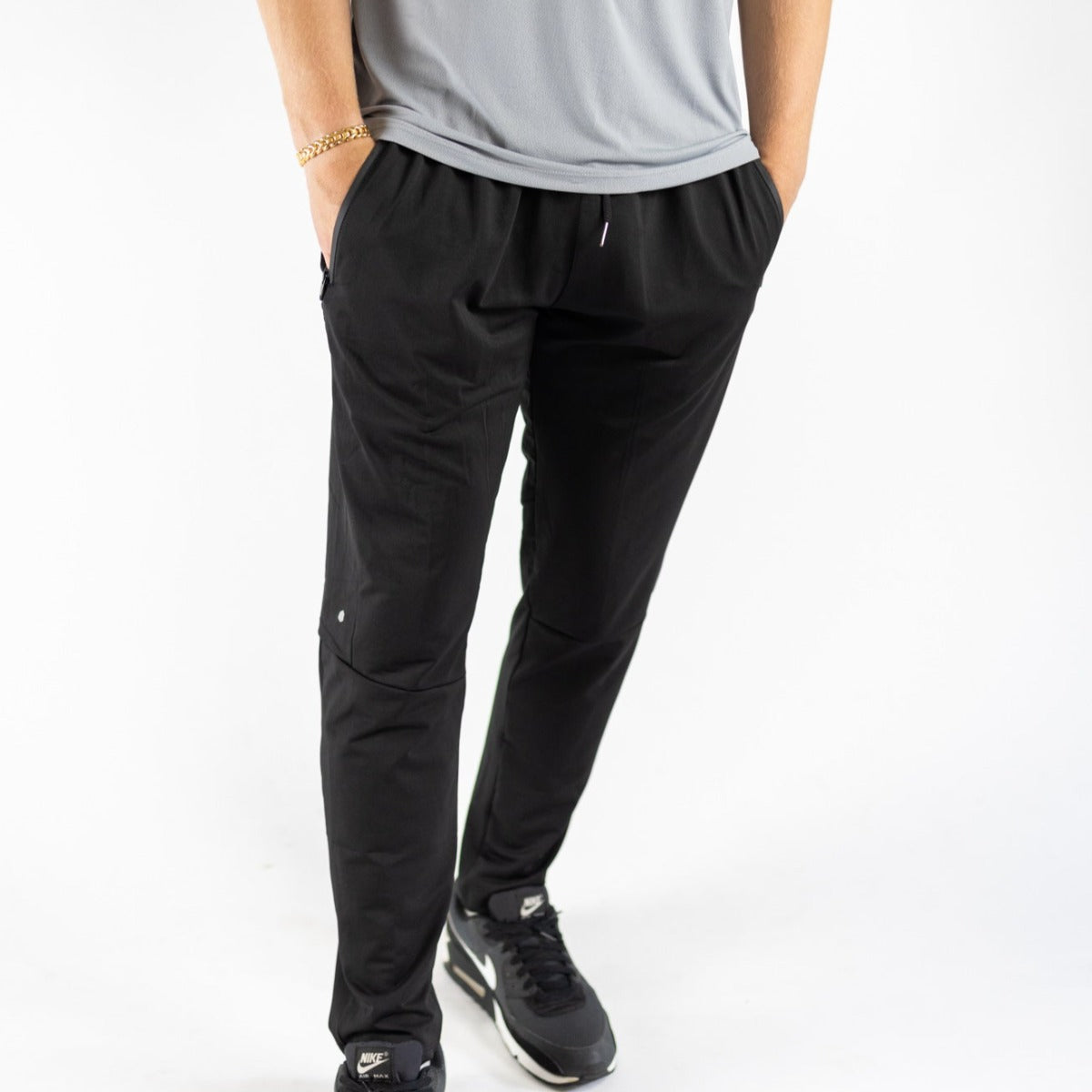A person wears DYM Athletics Men's Luxury Lounge Jogger in black, paired with Nike sneakers and a gold chain bracelet, standing with hands in pockets. Only the lower body is shown against a plain white background.