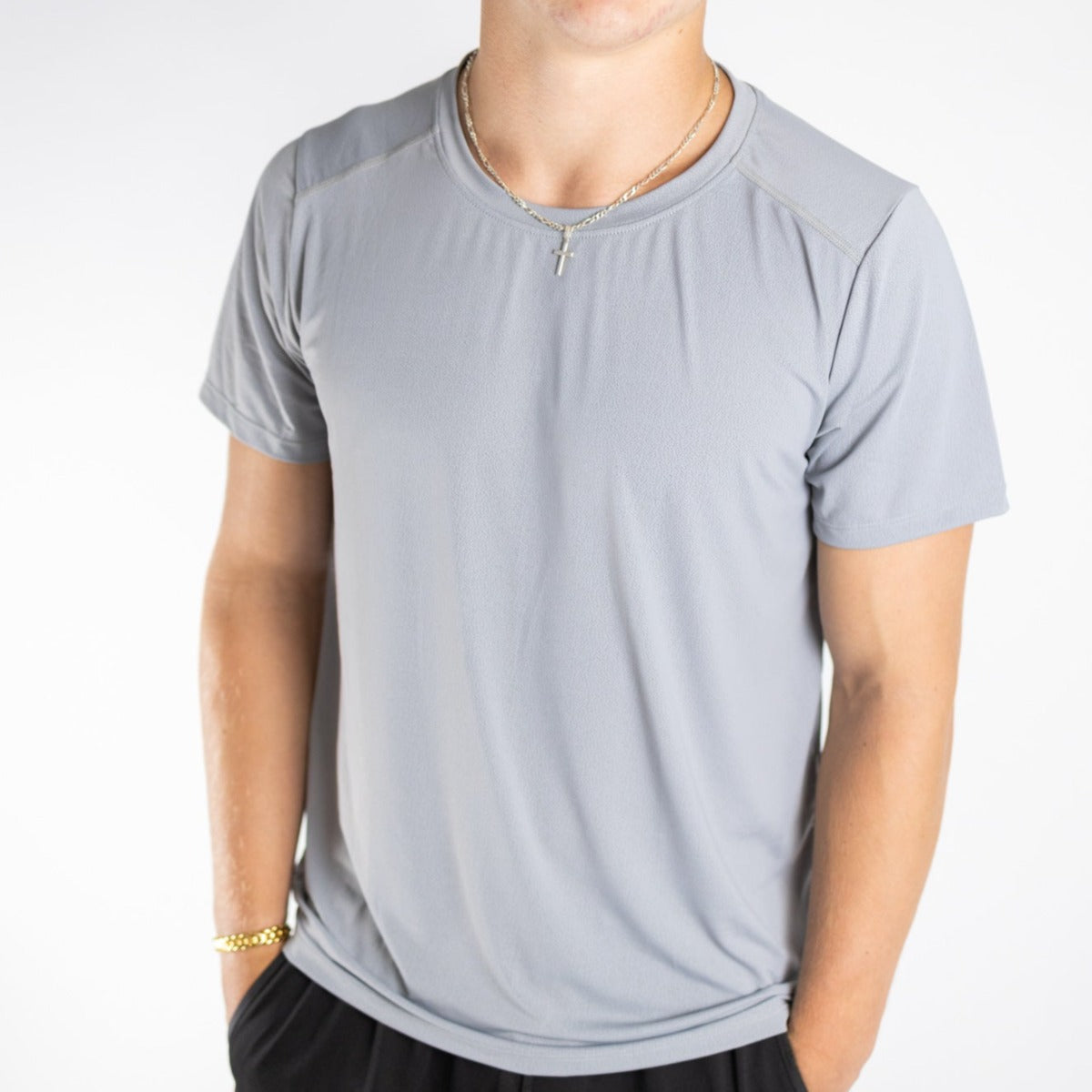 A person wears the DYM Athletics Elevate Men's Lightweight Breathable Tee in light gray and black pants, standing against a white background. They accessorize with a gold chain bracelet and cross necklace; their face is partially out of frame.