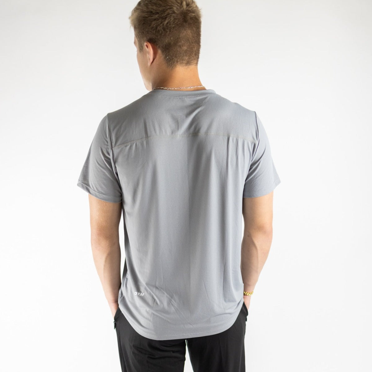 A person with short, light brown hair faces away from the camera, wearing a DYM Athletics Elevate Men's Lightweight Breathable Tee in light gray and black pants. Set against a plain white background, it’s ideal for casual wear.