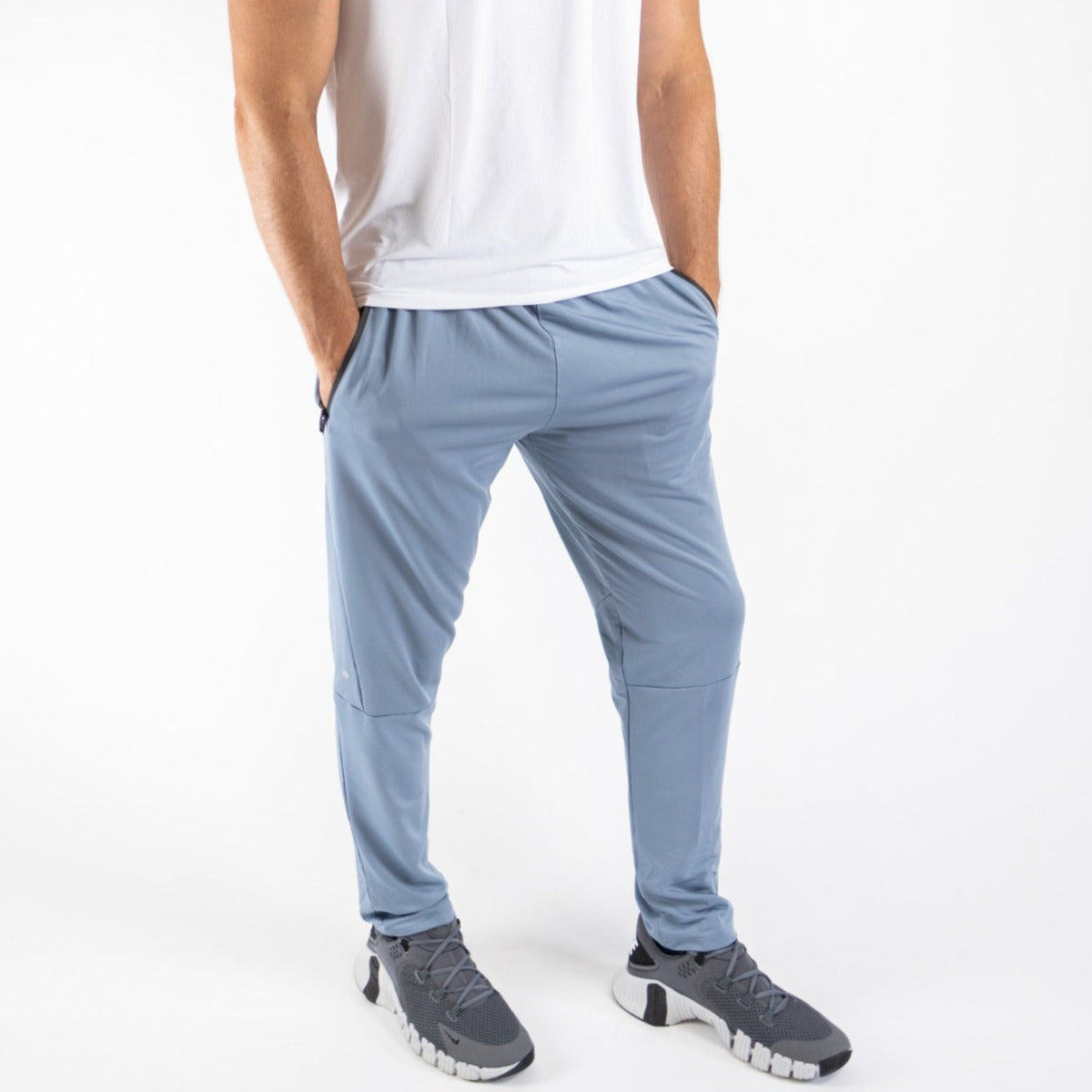 A person wears DYM Athletics Men's Luxury Lounge Jogger in light blue with zipper pockets and a white tee, paired with gray athletic shoes. Hands in pockets, standing against a white background; image is cropped at the shoulders.
