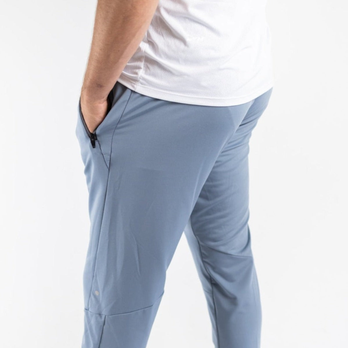 A person stands facing away, wearing DYM Athletics Men's Luxury Lounge Jogger | SALE in light blue with hands in pockets and a white t-shirt. The plain background highlights the fit and performance fabric of the pants.