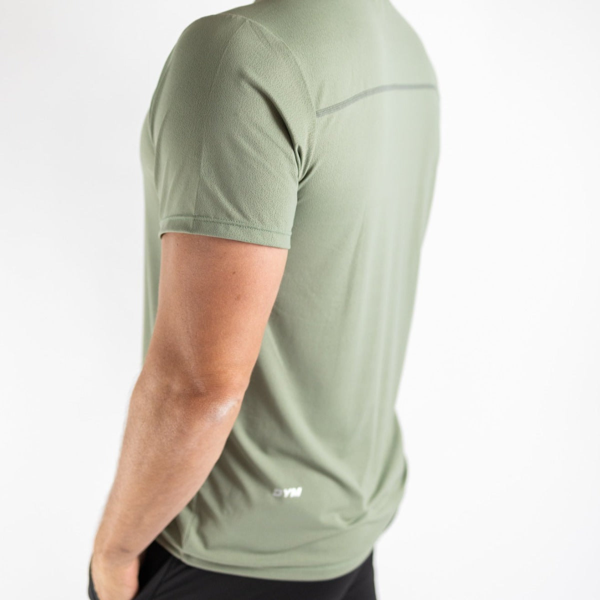 A man is seen from the side and back wearing the DYM Athletics Elevate Men's Lightweight Breathable Tee in light olive green with mesh panels, a thin upper-back seam, white hem logo, and black shorts—perfect for running or casual wear.