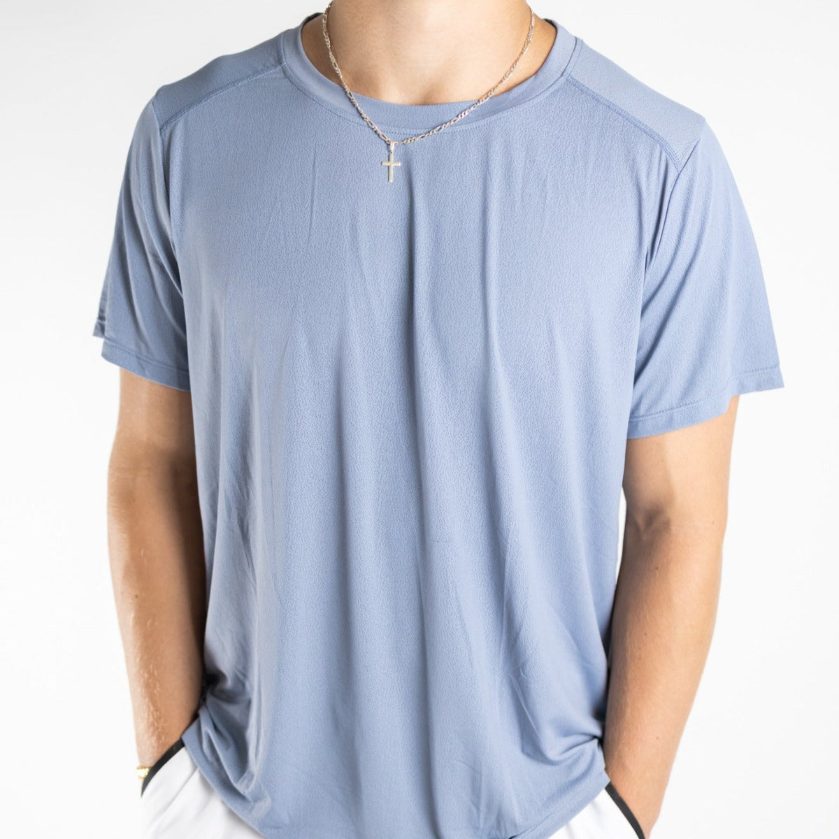 A man wears the DYM Athletics Elevate Men's Lightweight Breathable Tee in light blue, paired with a gold cross pendant and white pants—an ideal casual look, showcased against a plain white backdrop.