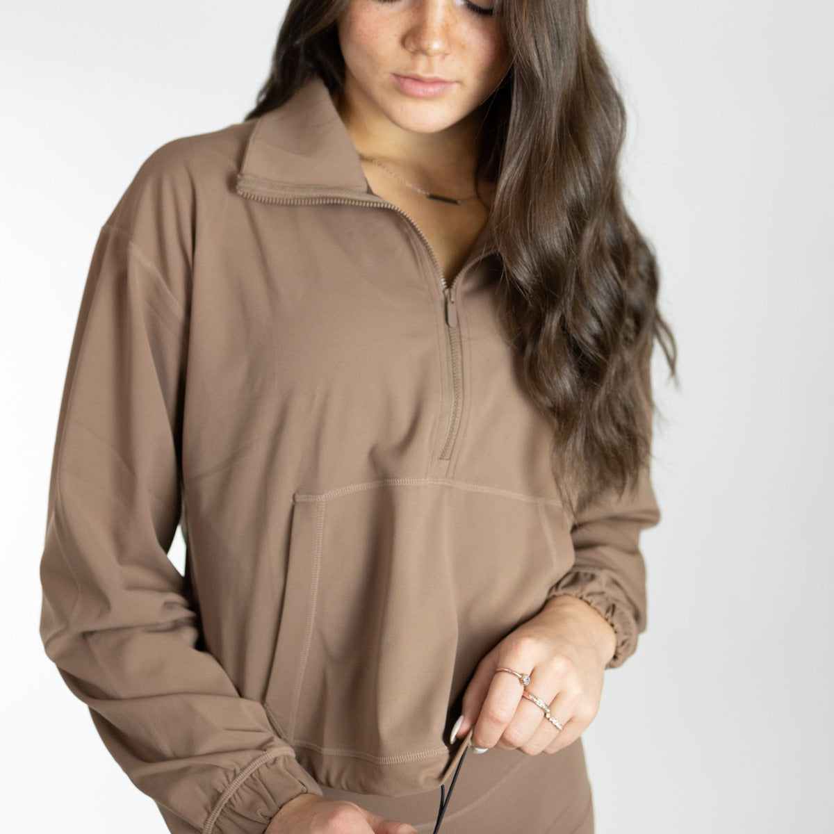 A woman with long brown wavy hair wears the DYM Athletics Ascend 1/4 Zip Jacket—a light brown, long-sleeve top with a kangaroo pocket and black drawstring waist. She looks down while adjusting it, accessorized with rings and a thin necklace.
