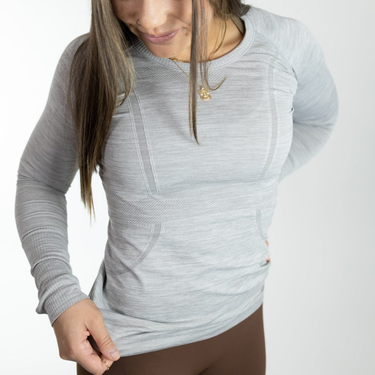 A person with long brown hair wears the DYM Athletics Trainer Lightweight Long Sleeve in light gray and brown pants against a white background. They look down, hold the shirt hem with one hand, and wear a gold pendant necklace.