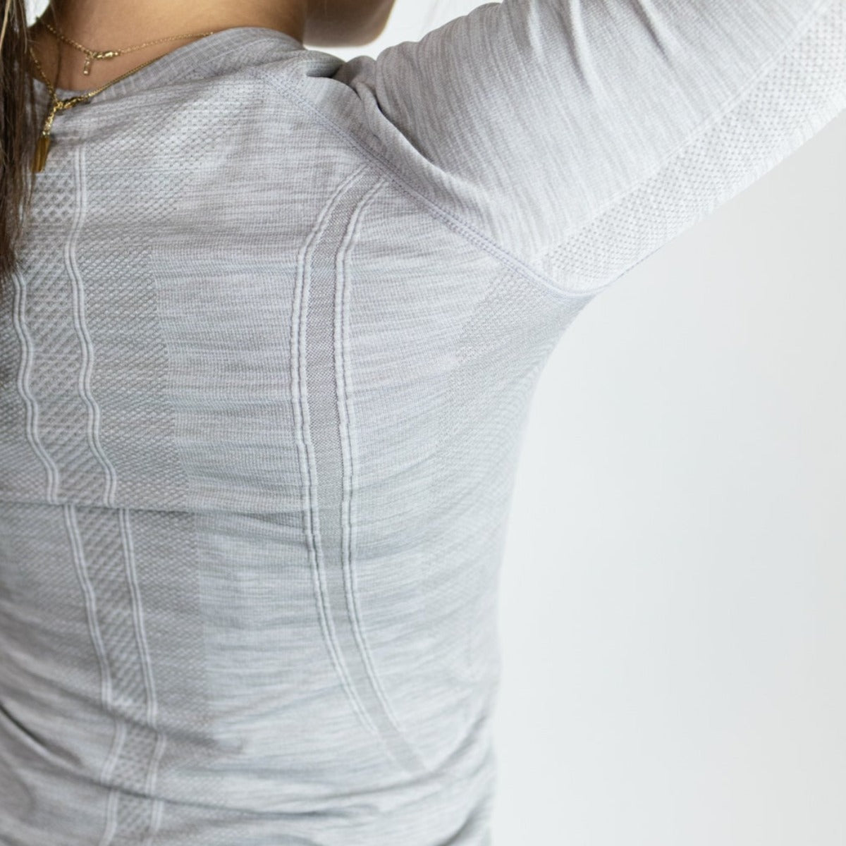 A close-up shows a person wearing the DYM Athletics Trainer Lightweight Long Sleeve in light gray. The sweat-wicking, textured fabric and fitted design with vertical patterns are highlighted, along with part of their long brown hair and a delicate necklace.