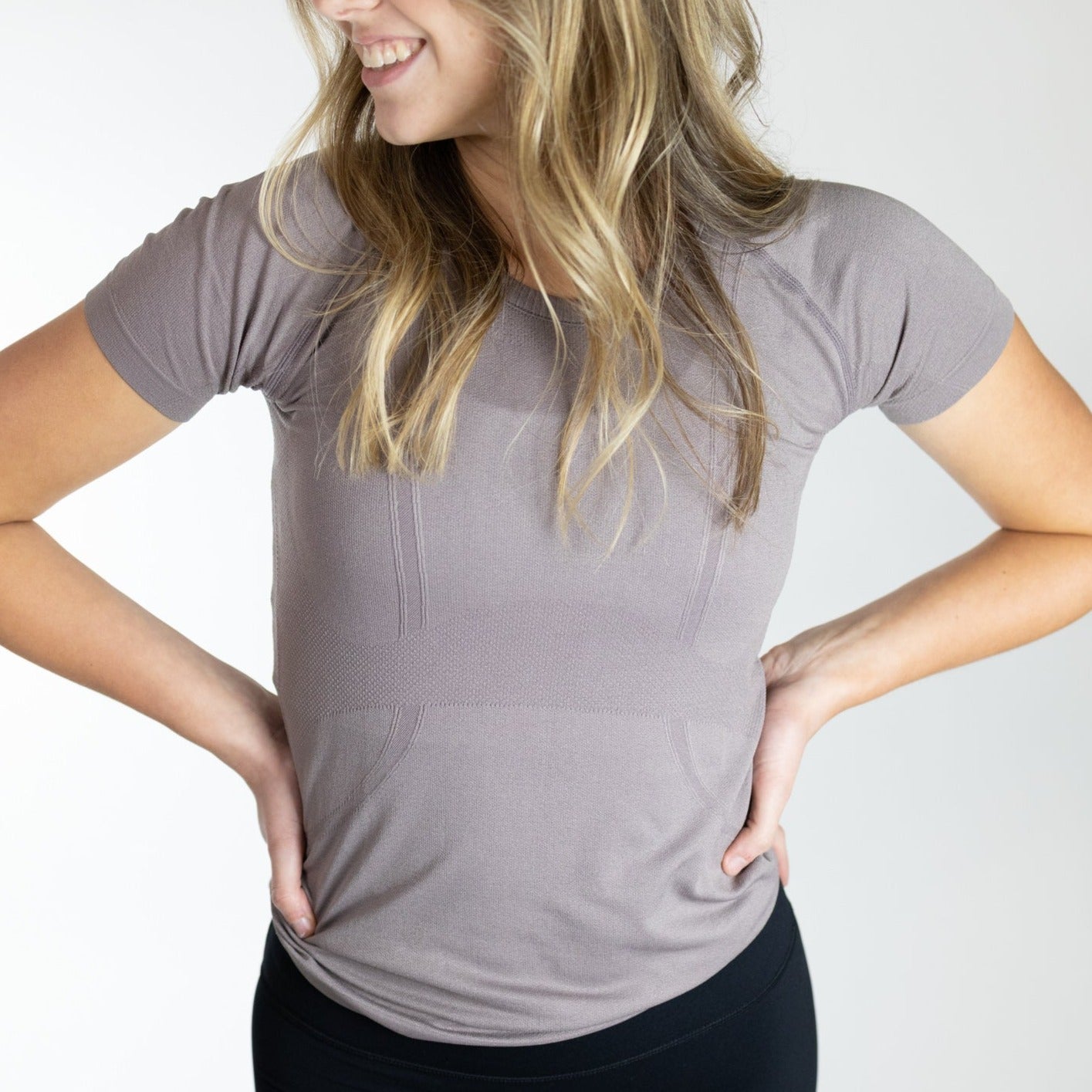 A woman with wavy blonde hair smiles, hands on hips, wearing DYM Athletics' Trainer Lightweight Short Sleeve Shirt in taupe and black leggings. She stands against a plain white background, her face partially turned to the side.