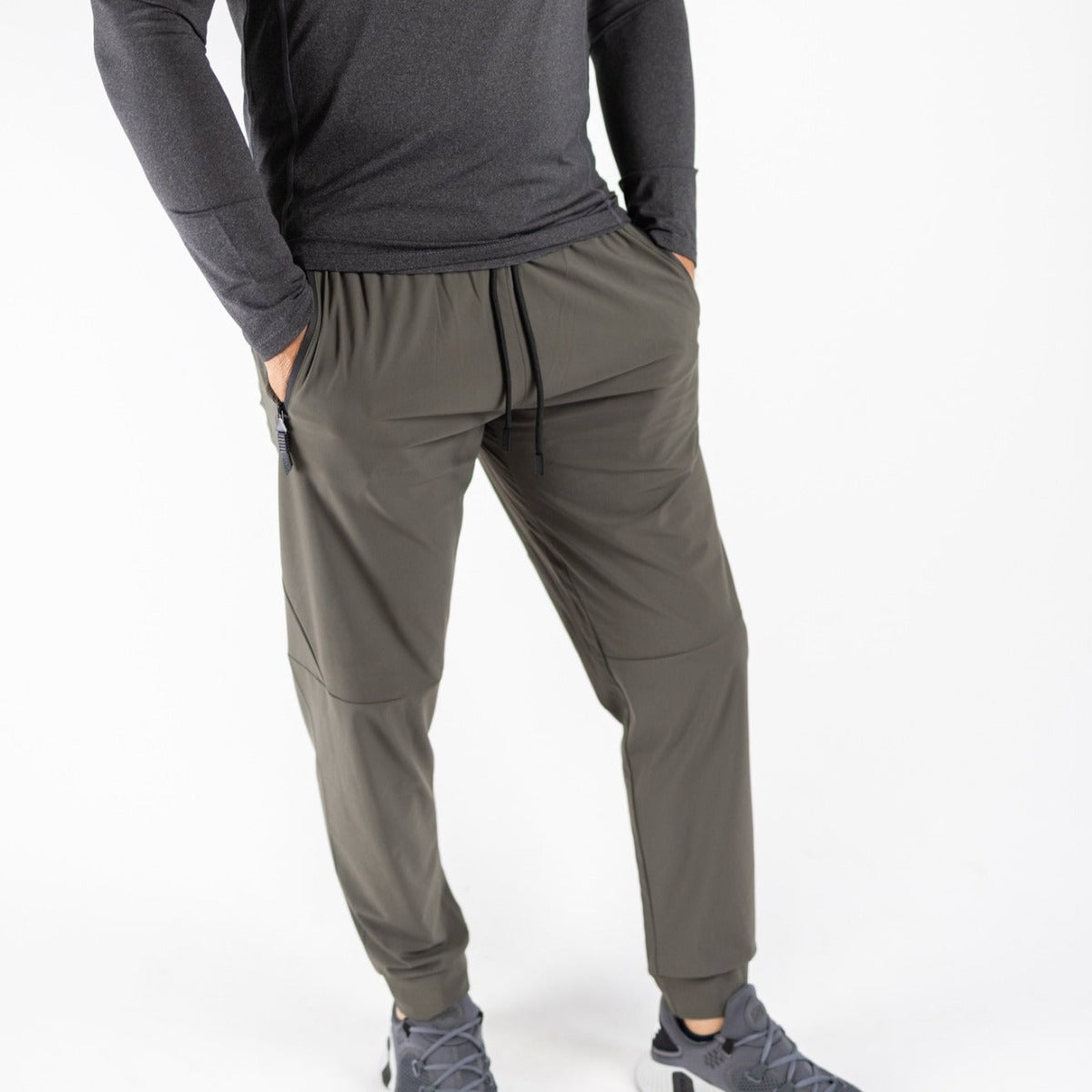 A person stands against a white background wearing DYM Athletics’ Fly Lightweight Active Jogger in olive green, featuring a drawstring waist and zipper pockets. They pair it with a dark long-sleeve shirt and gray athletic shoes. Face is not visible.