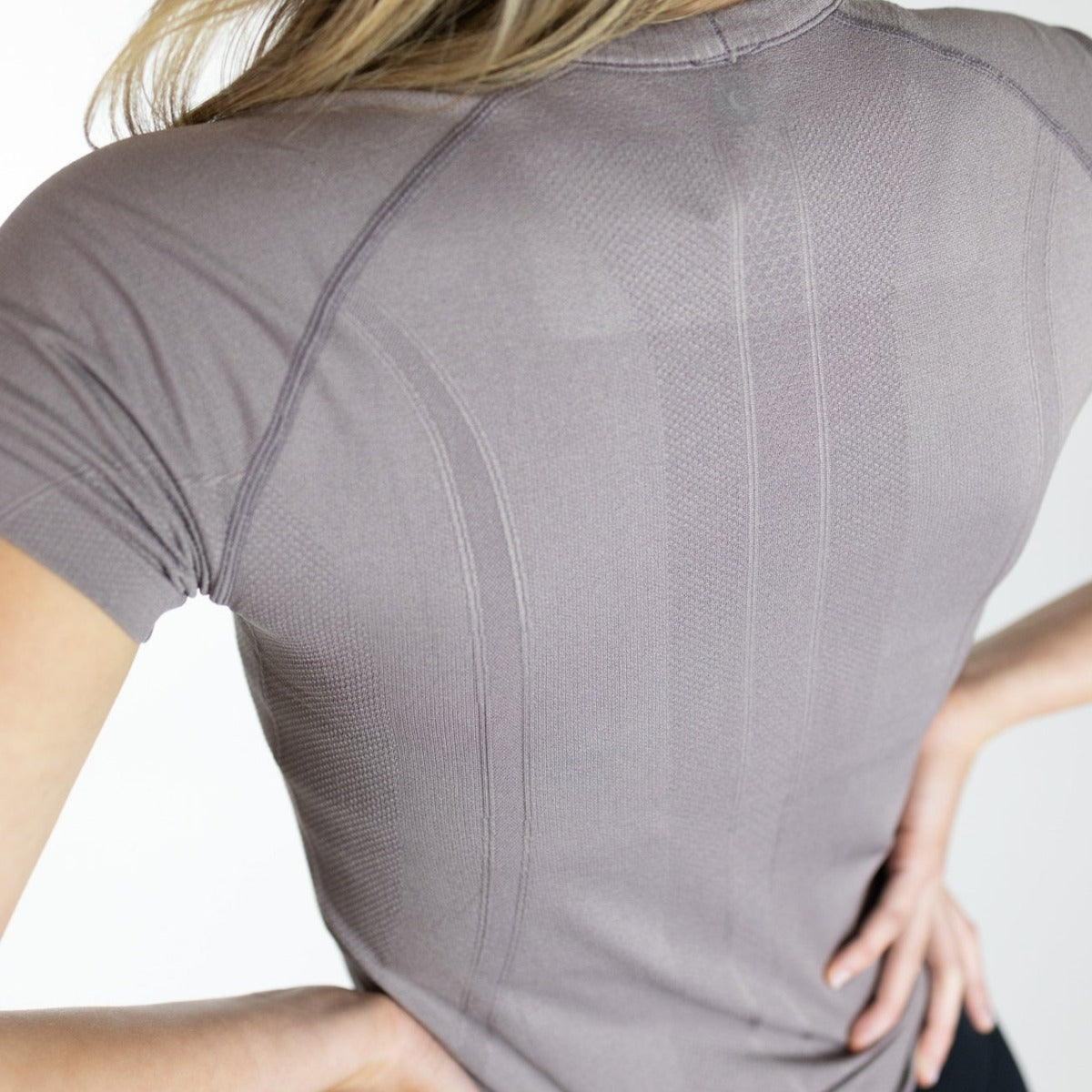 A person with long blonde hair stands with hands on hips, wearing the DYM Athletics Trainer Lightweight Short Sleeve Shirt in gray. The fitted, sweat-wicking fabric features subtle textured patterns, shown against a plain white background.
