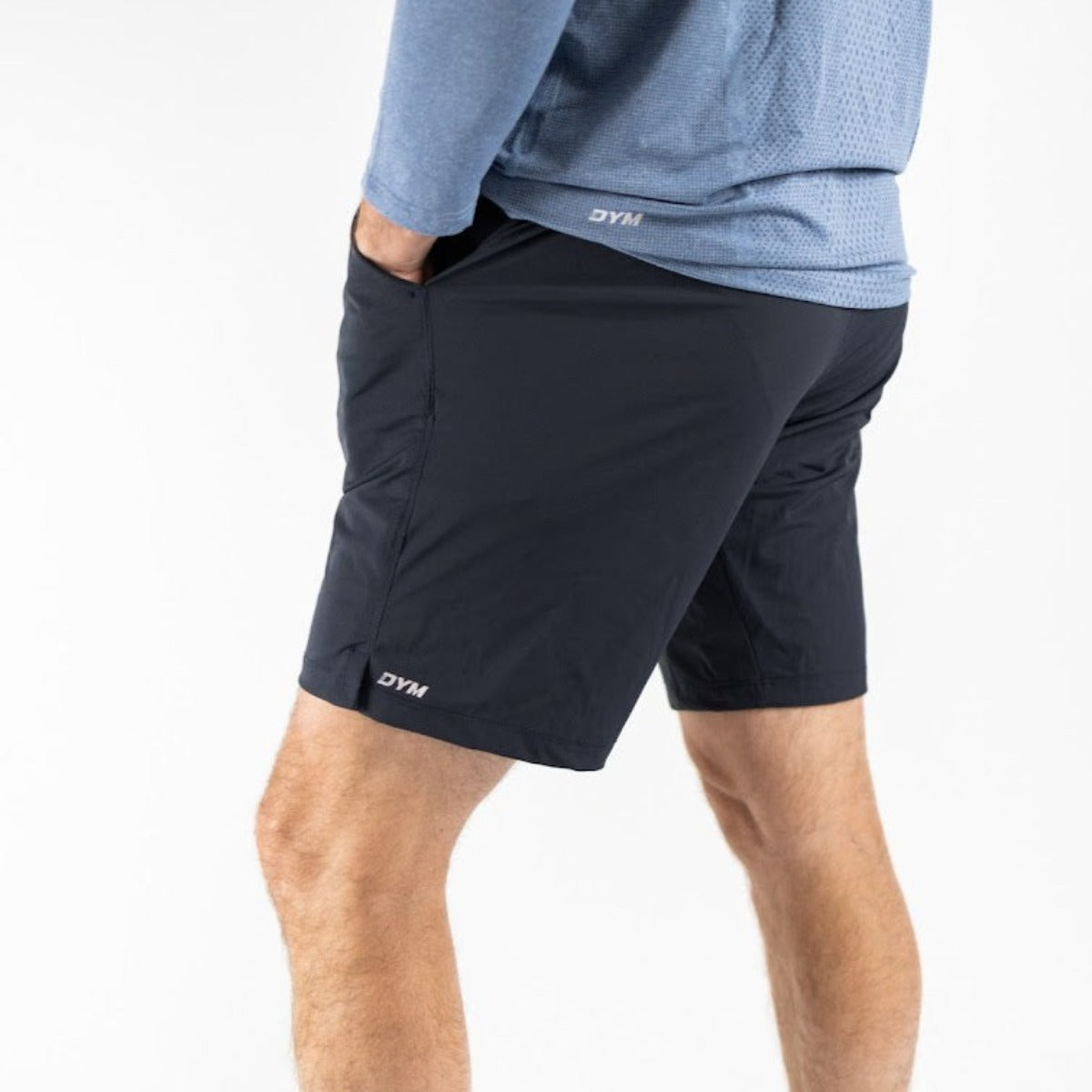 A person stands with hands in pockets, wearing a blue long-sleeve shirt and black DYM Athletics Active Moisture Wicking Short - 9". "DYM" is printed near the hem. Only the lower torso and legs are visible against a white background.