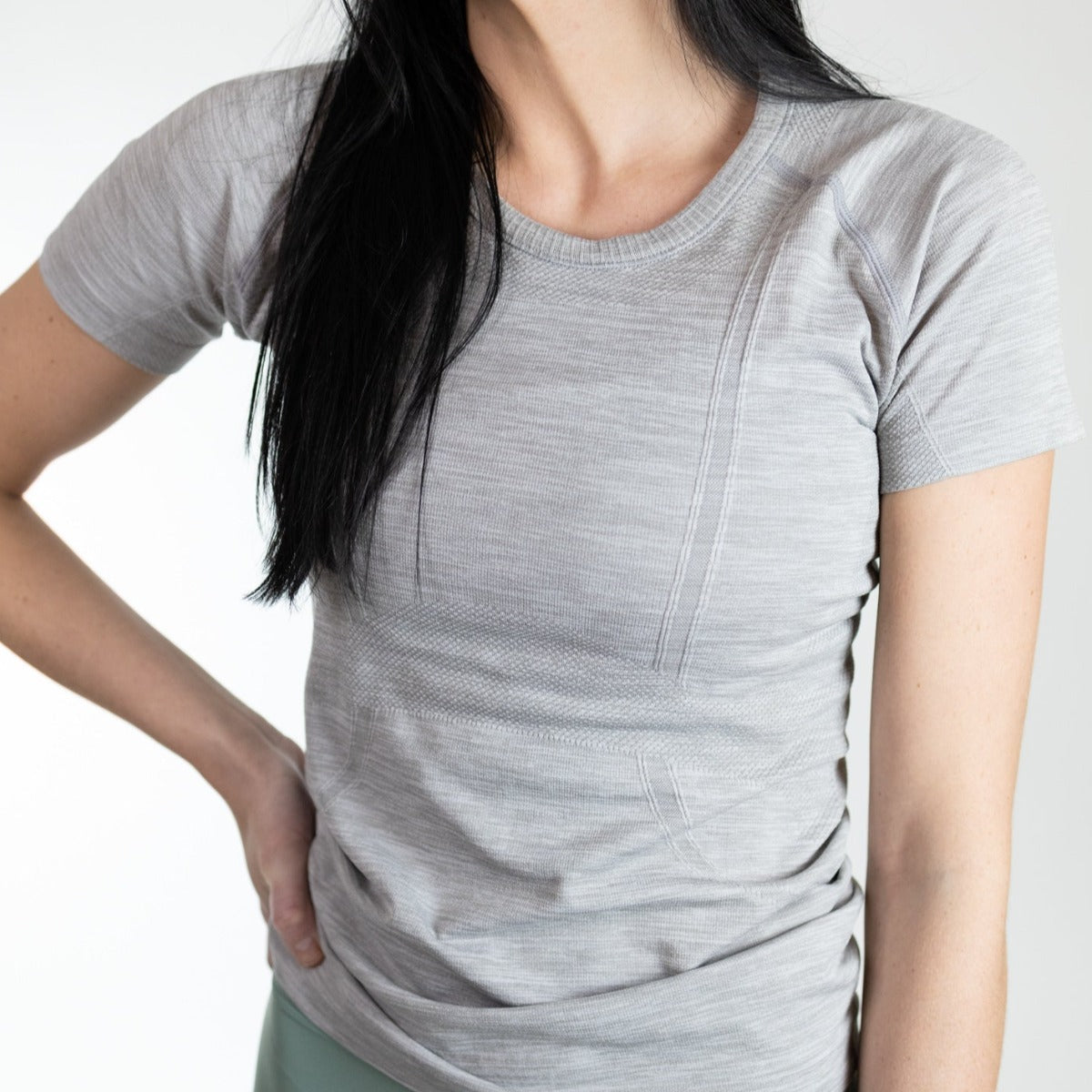 A woman with long black hair models the DYM Athletics Trainer Lightweight Short Sleeve Shirt in light gray, standing with one hand on her hip against a plain white background. Her face is not visible.
