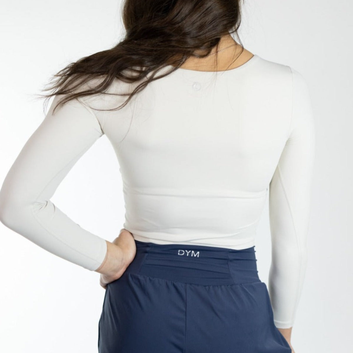 A person with long brown hair stands facing away from the camera, wearing a fitted Flex Long Sleeve w/ Built in Bra by DYM Athletics and high-waisted dark blue athletic pants with a small DYM logo. Their left hand rests on their hip against a plain white background.