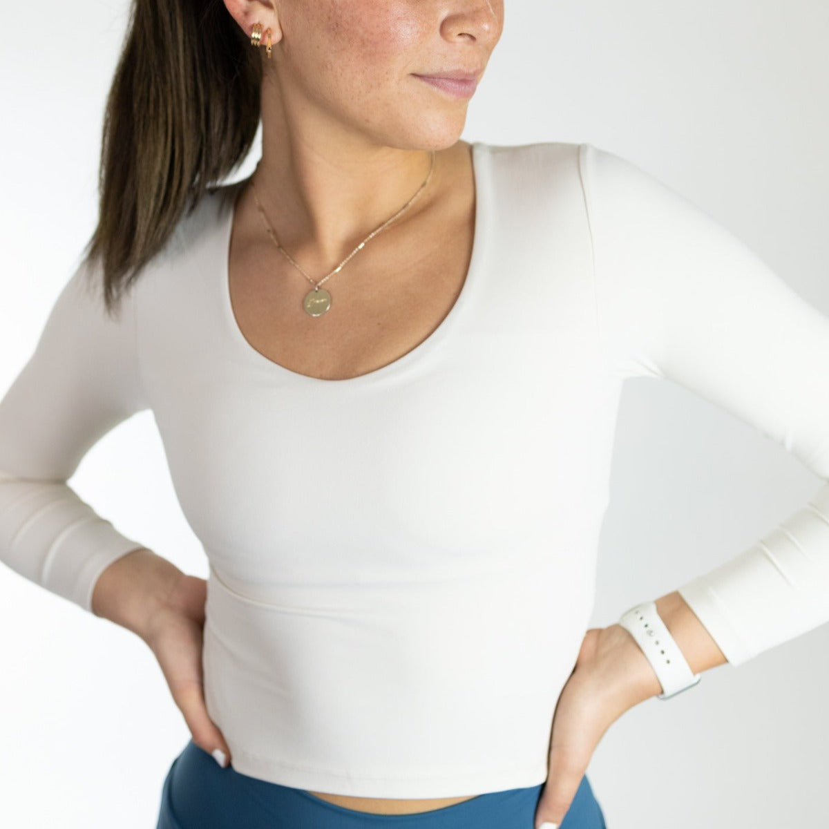 A woman stands with hands on hips, wearing a fitted white Flex Long Sleeve w/ Built in Bra by DYM Athletics and blue high-waisted leggings. She accessorizes with a gold necklace, hoop earrings, white watch, and has brown hair tied back while looking to the side.