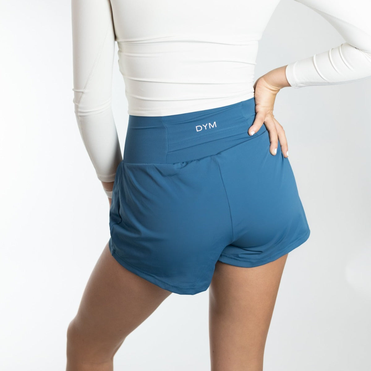 A person is shown from the waist down, wearing a fitted long-sleeve white top and DYM Athletics Namaste Yoga Shorts | 3" with "DYM" on the waistband. One hand rests on their hip against a minimalistic white background.