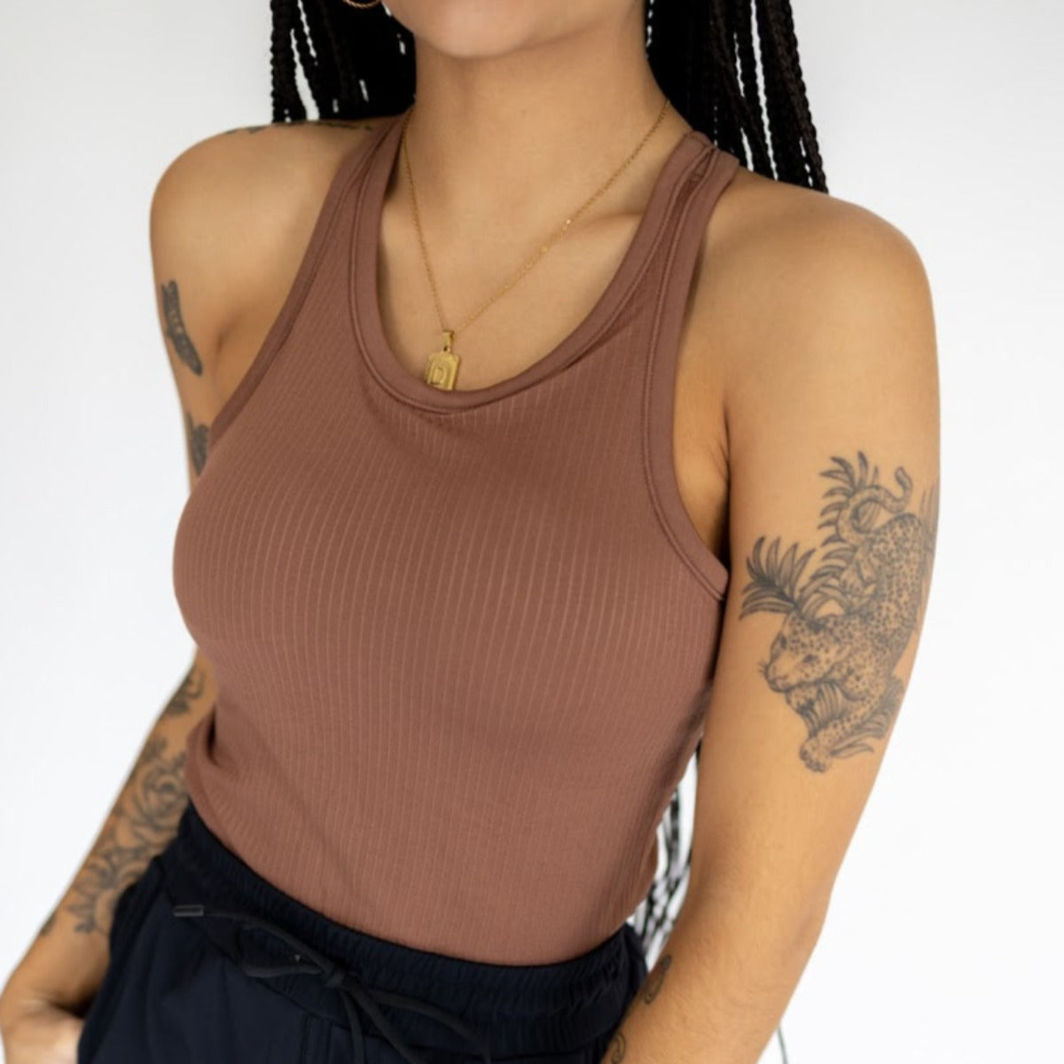 A woman with long braids wears the DYM Athletics Restore Ribbed Comfort Tank w/ Built-in Bra and dark pants, accessorized with a gold necklace. She has tattoos, including a leopard and foliage on her right arm, against a plain white background.