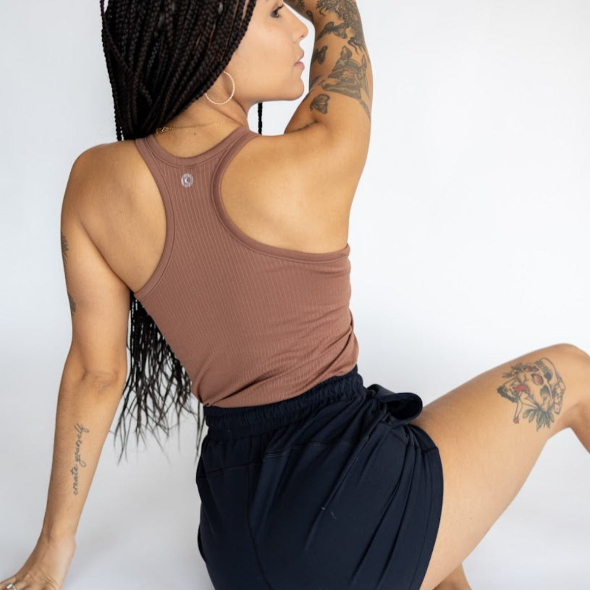 A person with long braided hair sits on the floor, facing away, wearing the DYM Athletics Restore Ribbed Comfort Tank w/ Built in Bra in brown and black shorts. Tattoos adorn their left arm and right thigh; a plain white background completes the scene.