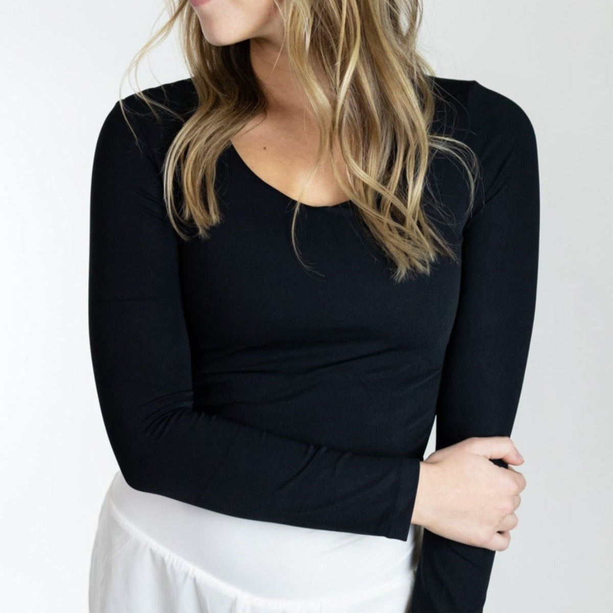 A woman with wavy blonde hair, partially turned sideways, wears a fitted black DYM Athletics Flex Long Sleeve w/ Built in Bra and a white skirt. Her left arm holds her right elbow. The background is plain and light-colored; her face is partly out of view.