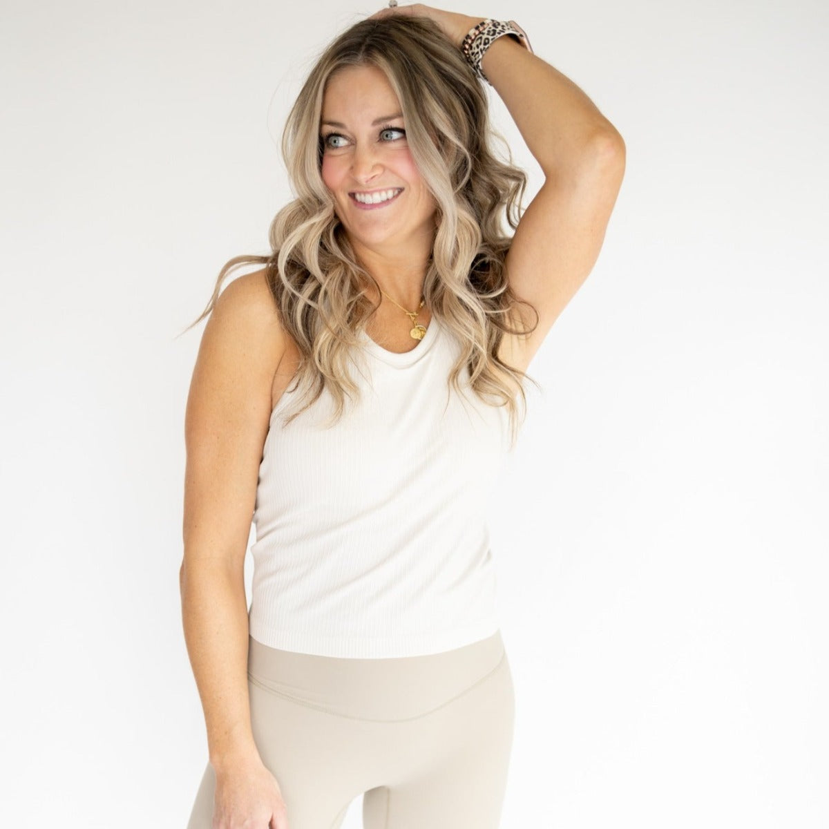 A woman with wavy blonde hair smiles to her left, wearing the DYM Athletics Restore Ribbed Comfort Tank w/ Built in Bra and beige leggings. She poses with one hand above her head against a clean, white background.