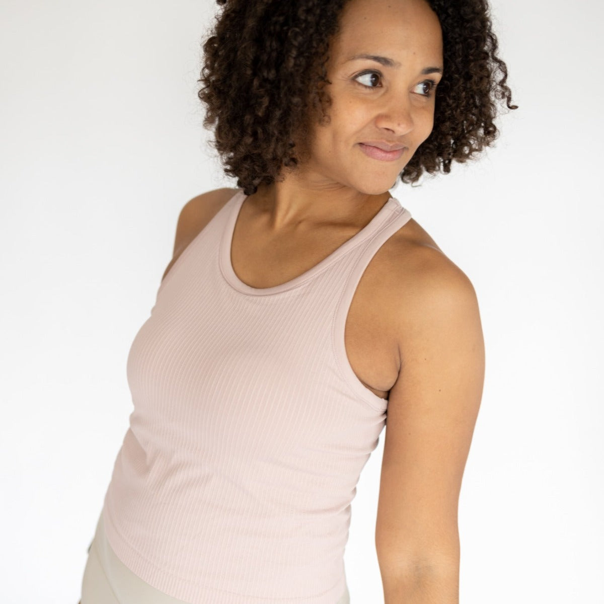 A woman with medium brown curly hair models the DYM Athletics Restore Ribbed Comfort Tank w/ Built in Bra in light pink with light pants, standing against a white background, smiling gently and looking left with her body slightly angled away.