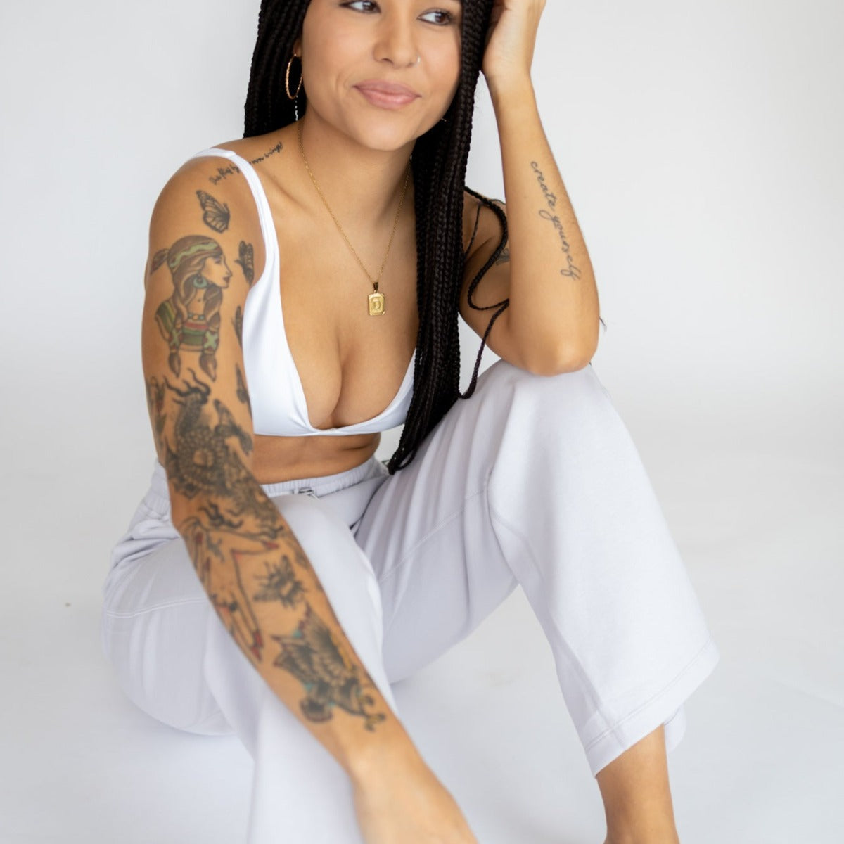 A woman with long braids and visible arm tattoos sits barefoot on the floor, smiling softly as she wears a white crop top and DYM Athletics Relax Wide Leg Sweatpant w/ Cinch Drawstring, set against a plain white background.