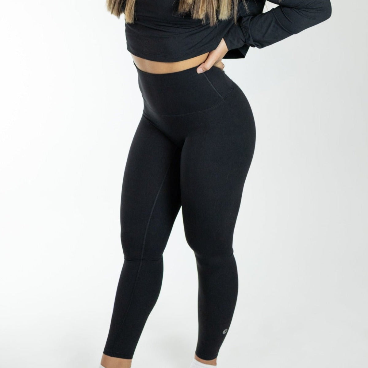 A woman with long, highlighted hair poses against a white background wearing DYM Athletics' Ultra High Waist Flex & Flow Legging and matching black crop top. Her left hand is on her hip, and her upper face is out of frame.