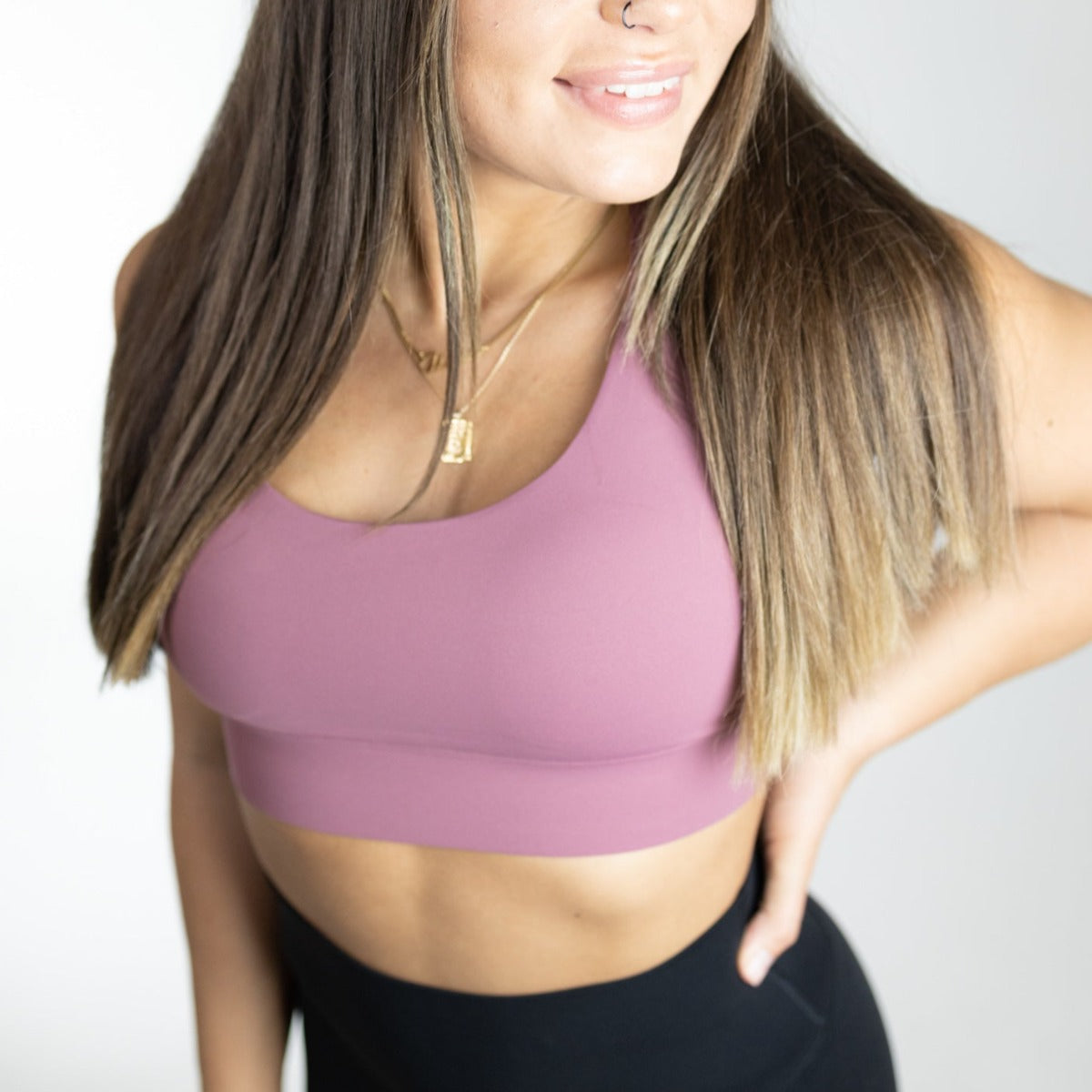 A woman with long brown hair and a nose ring smiles in the mauve DYM Athletics Impact Bra for A-D+ cup (High Impact), paired with black leggings, accessorized with gold necklaces, against a plain light background.