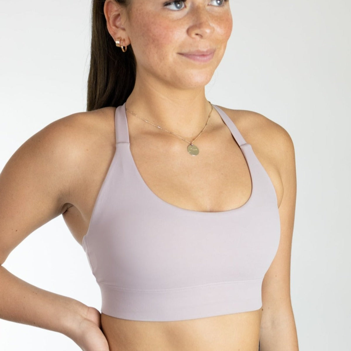 A fair-skinned, freckled woman wears the DYM Athletics Impact Bra | A-D+ cup in light purple, made from buttery soft fabric, paired with a gold pendant necklace and small hoop earrings. Her brown hair is pulled back as she smiles against a plain background.