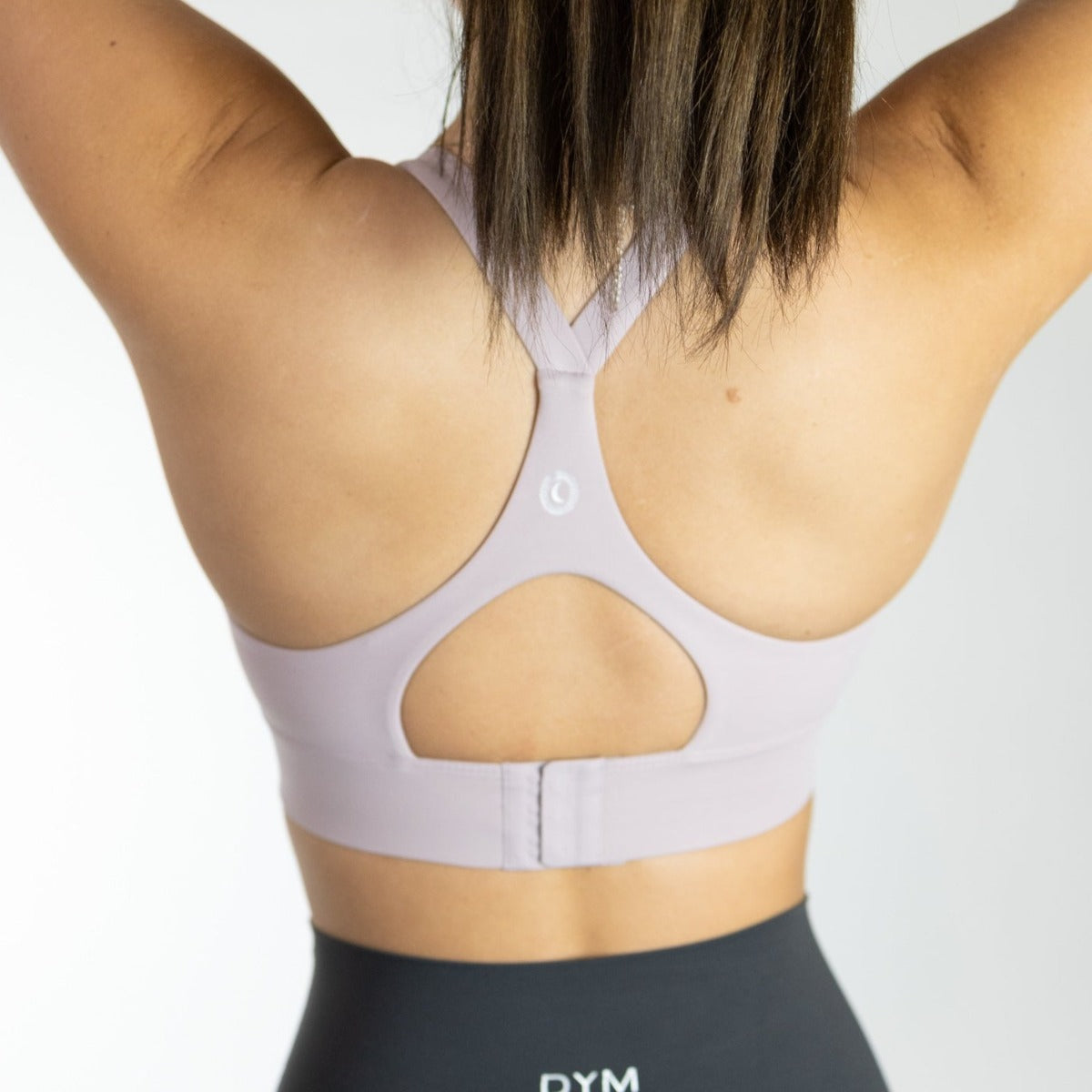 A woman, shown from behind with arms raised, wears the DYM Athletics Impact Bra in light purple (A-D+ cup, high impact) and black leggings. The buttery soft fabric stands out against a white background as her straight brown hair falls to her shoulders.