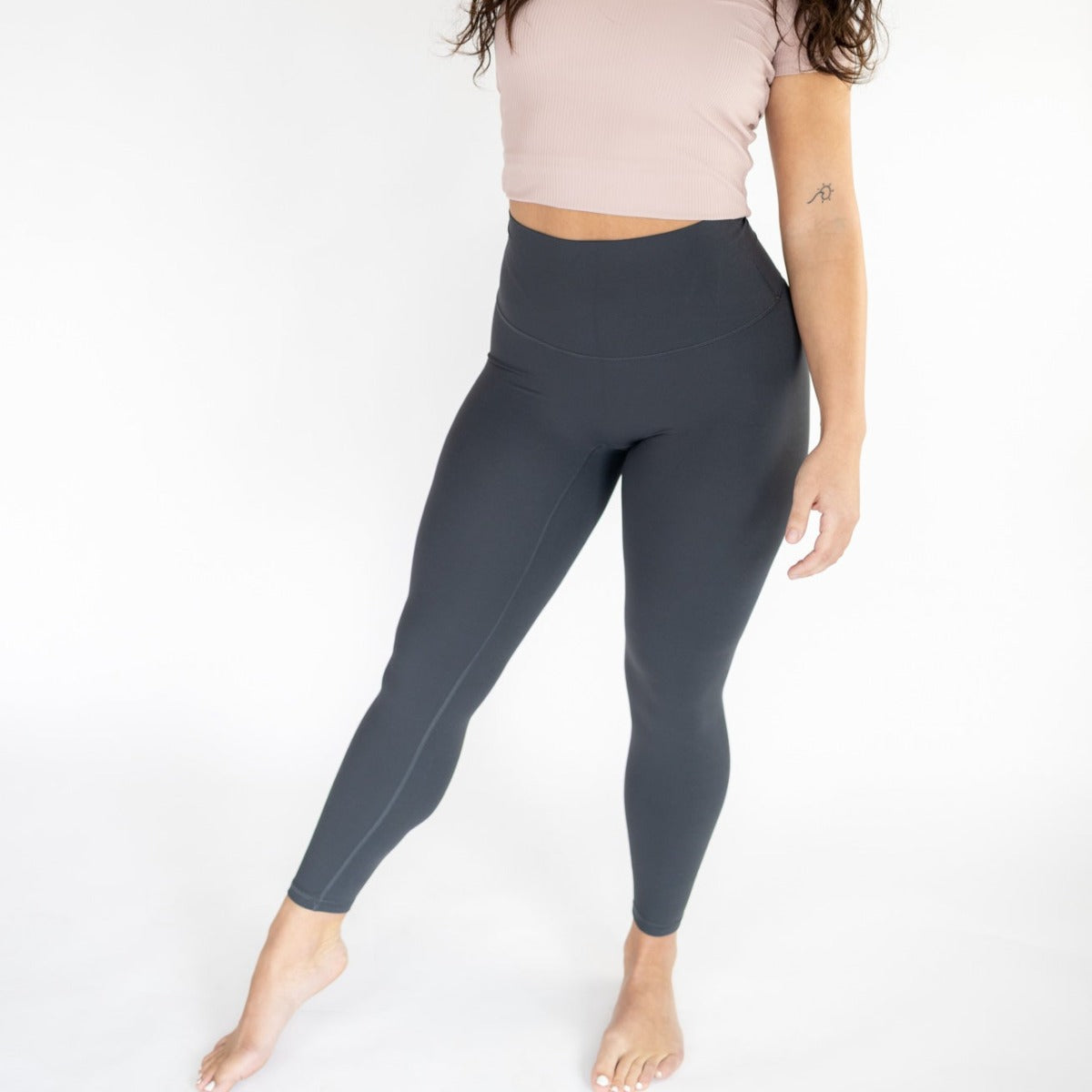 A person stands barefoot on a white background, wearing DYM Athletics Ultra High Waist Flex & Flow Legging in dark gray and a cropped light pink short-sleeve top. Their head is partially out of frame; one leg is bent, showing a tattoo on the left upper arm.