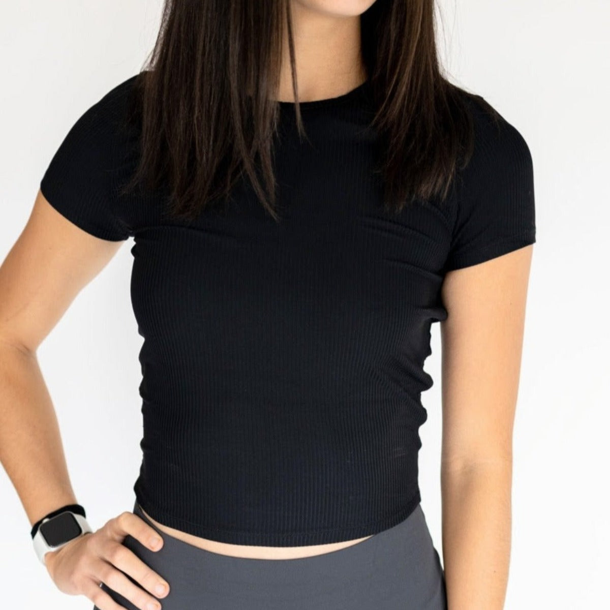 A woman with straight brown hair wears the DYM Athletics Restore Ribbed T-shirt in black and dark high-waisted leggings, standing against a white background. She rests her left hand with a smart watch on her hip.
